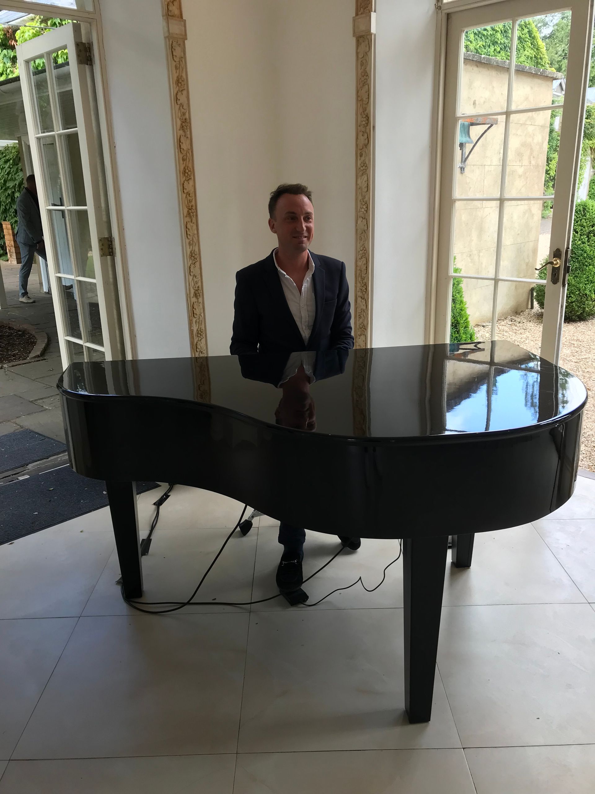 A man in a white shirt with an open collar and a blue blazer sits in front of a black baby grand piano 