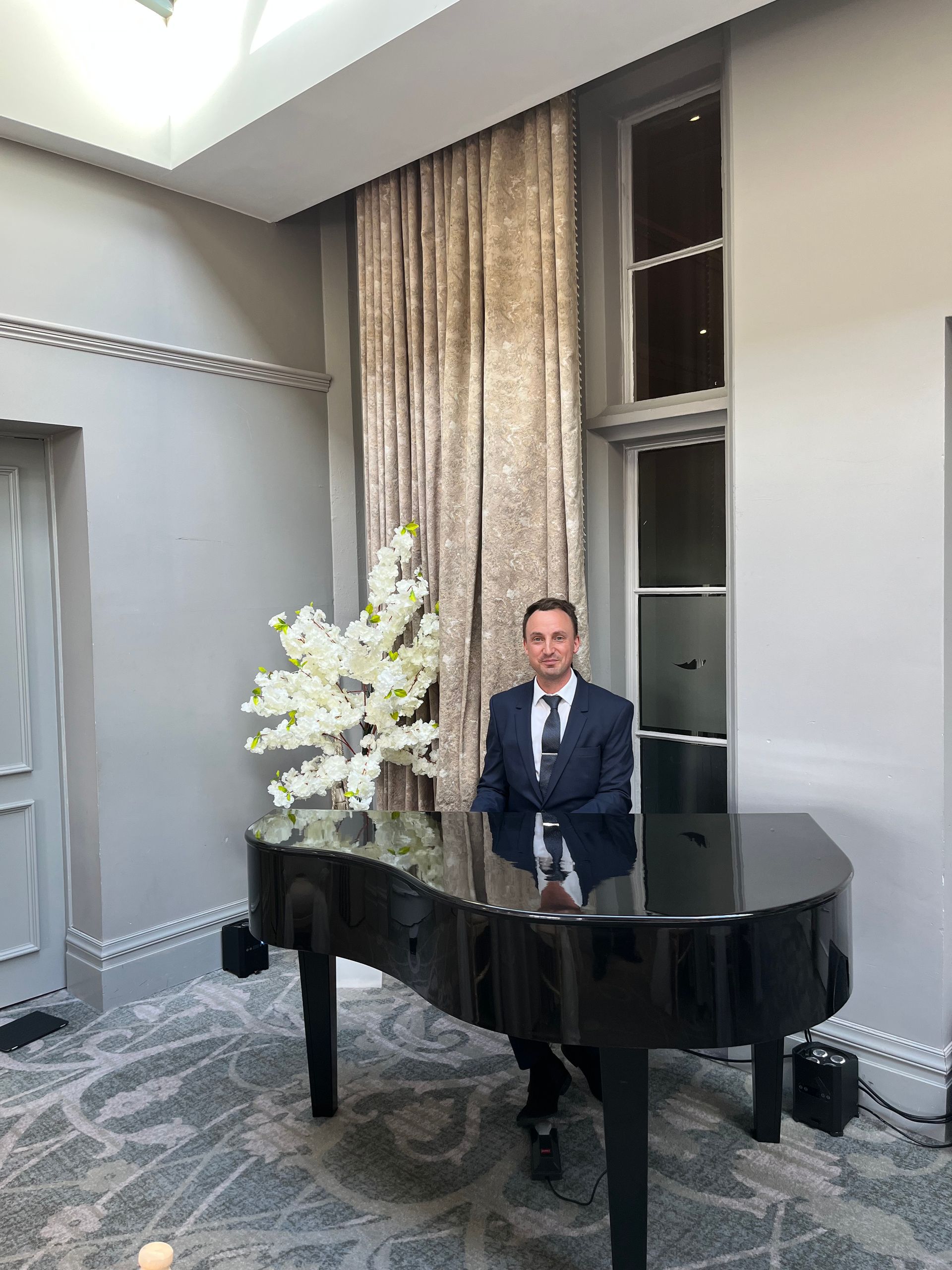 A man in a suits sits at a black grand piano in front of a window and next to some white flowers
