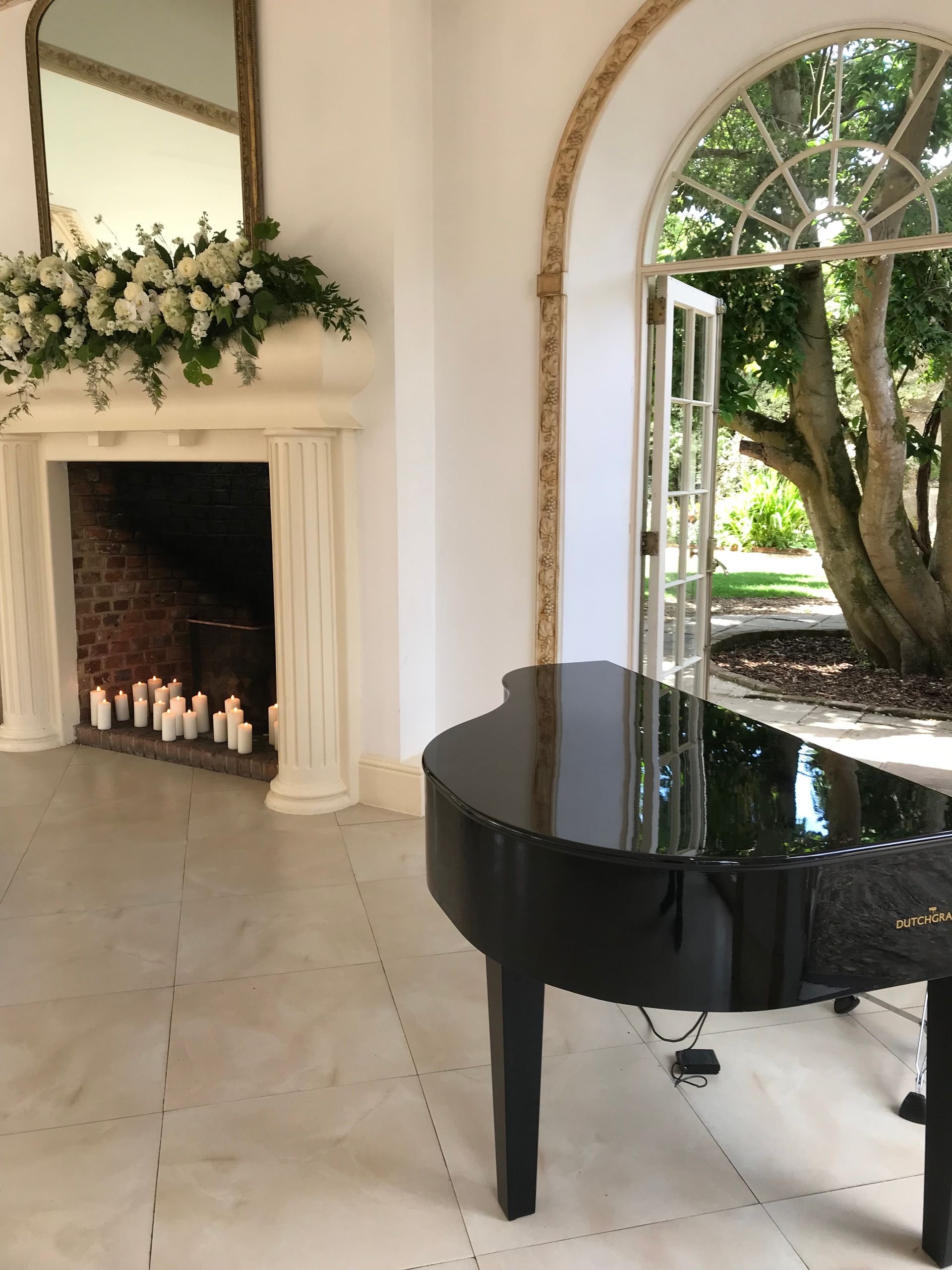 A black grand piano is in front of a fireplace with candles whilst French doors are wide open looking out into the outside