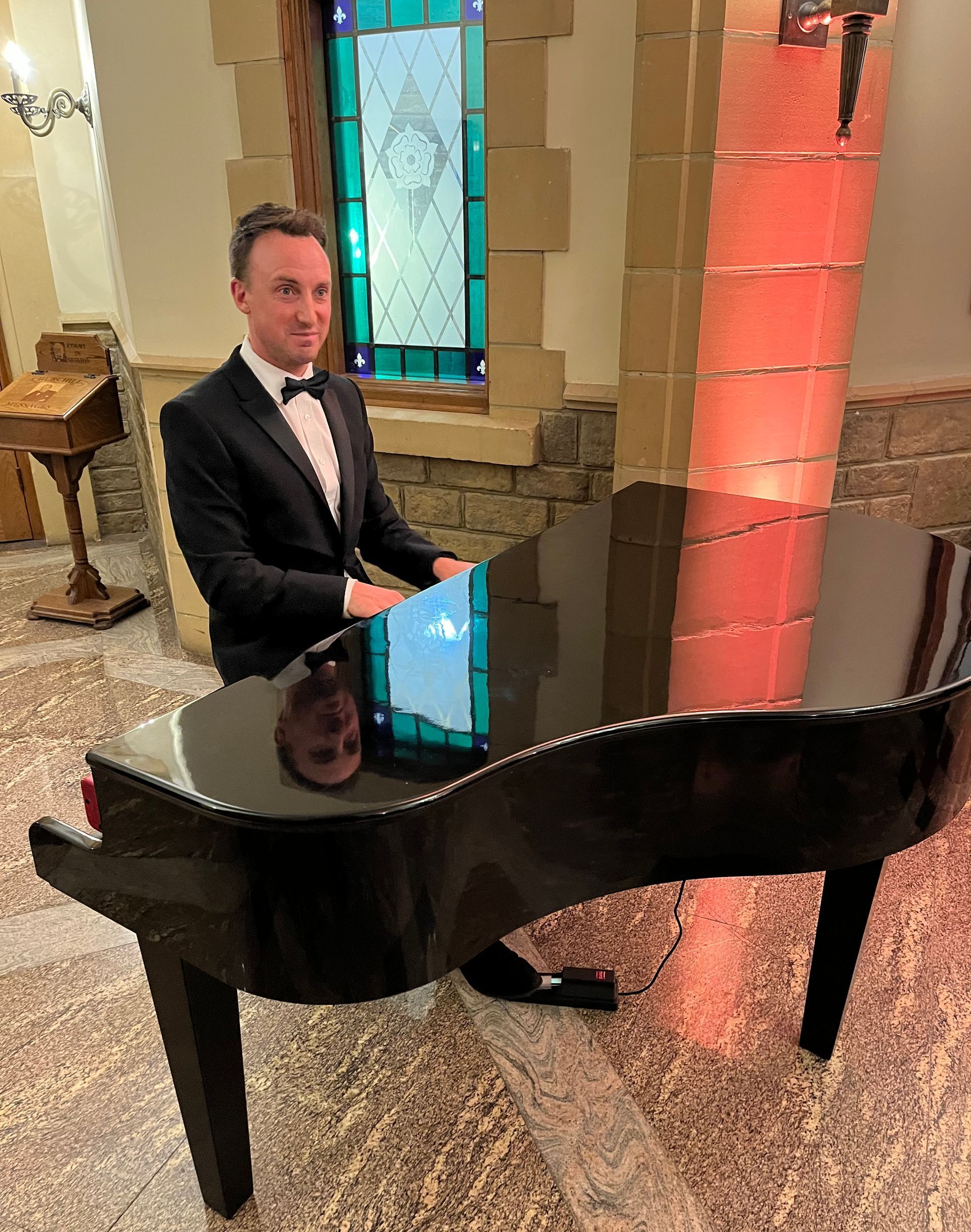 A man in a tuxedo is sitting at a piano in front of a stained glass window.