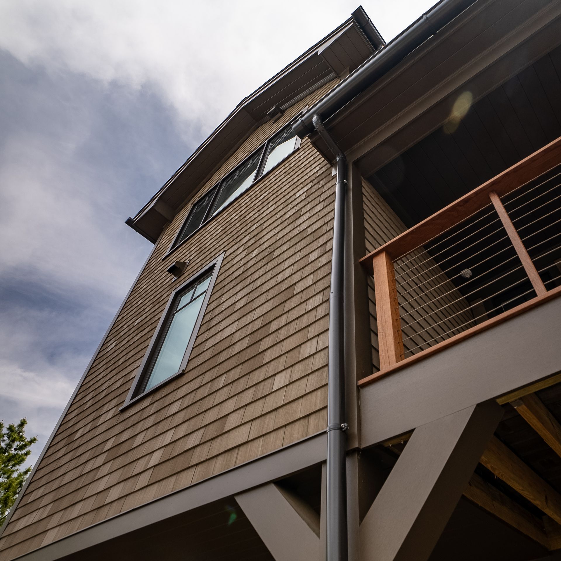 Upward view of a brown shingled building with windows, balcony railing, and cloudy sky