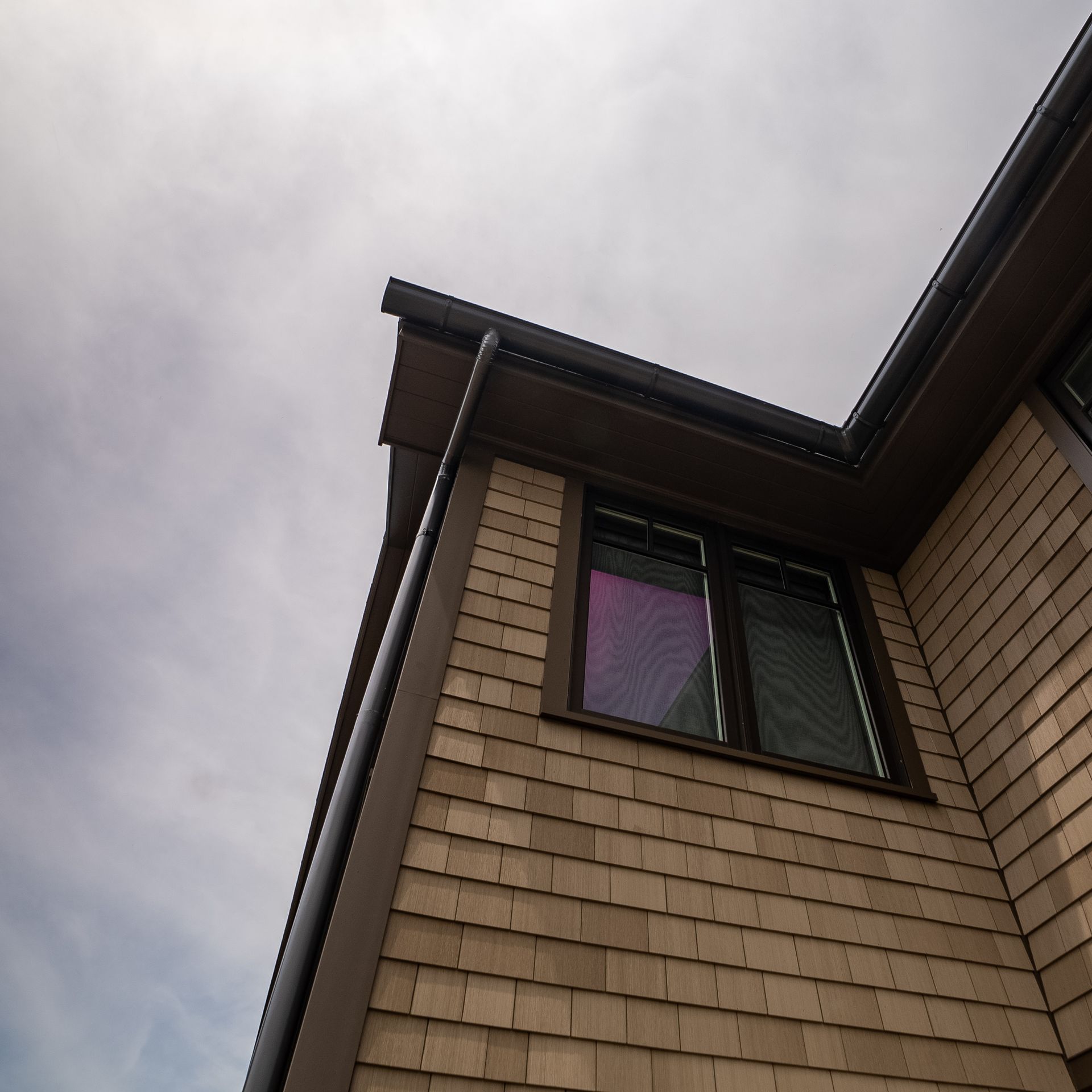 Corner of a beige shingled house with a dark roof and a window under a cloudy sky