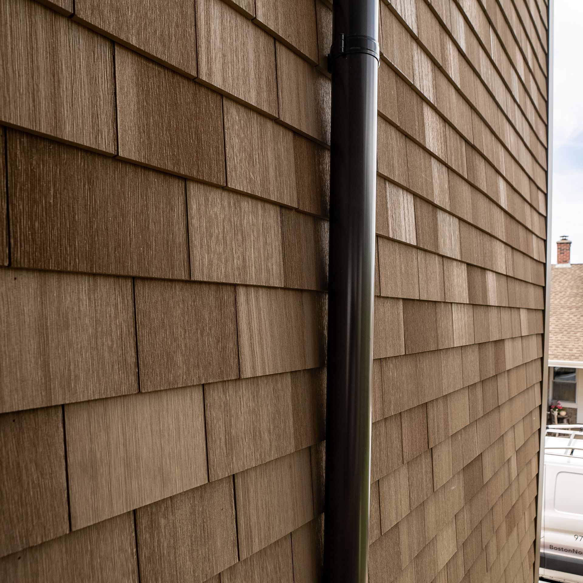 Brown shingle siding on a building corner with a black downspout and a glimpse of street below