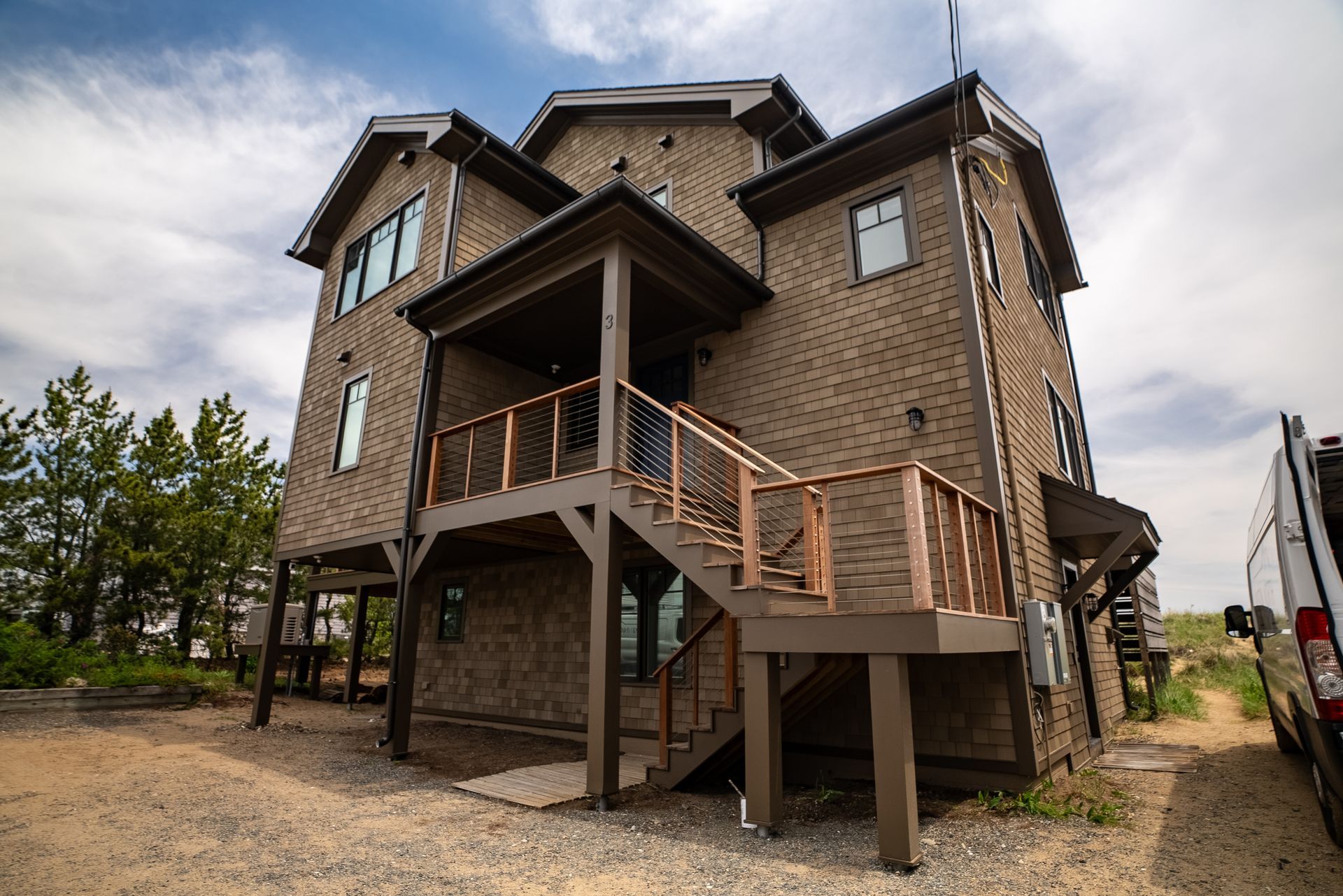 Elevated brown house with wraparound porch and wooden stairs on a gravel lot under a cloudy sky
