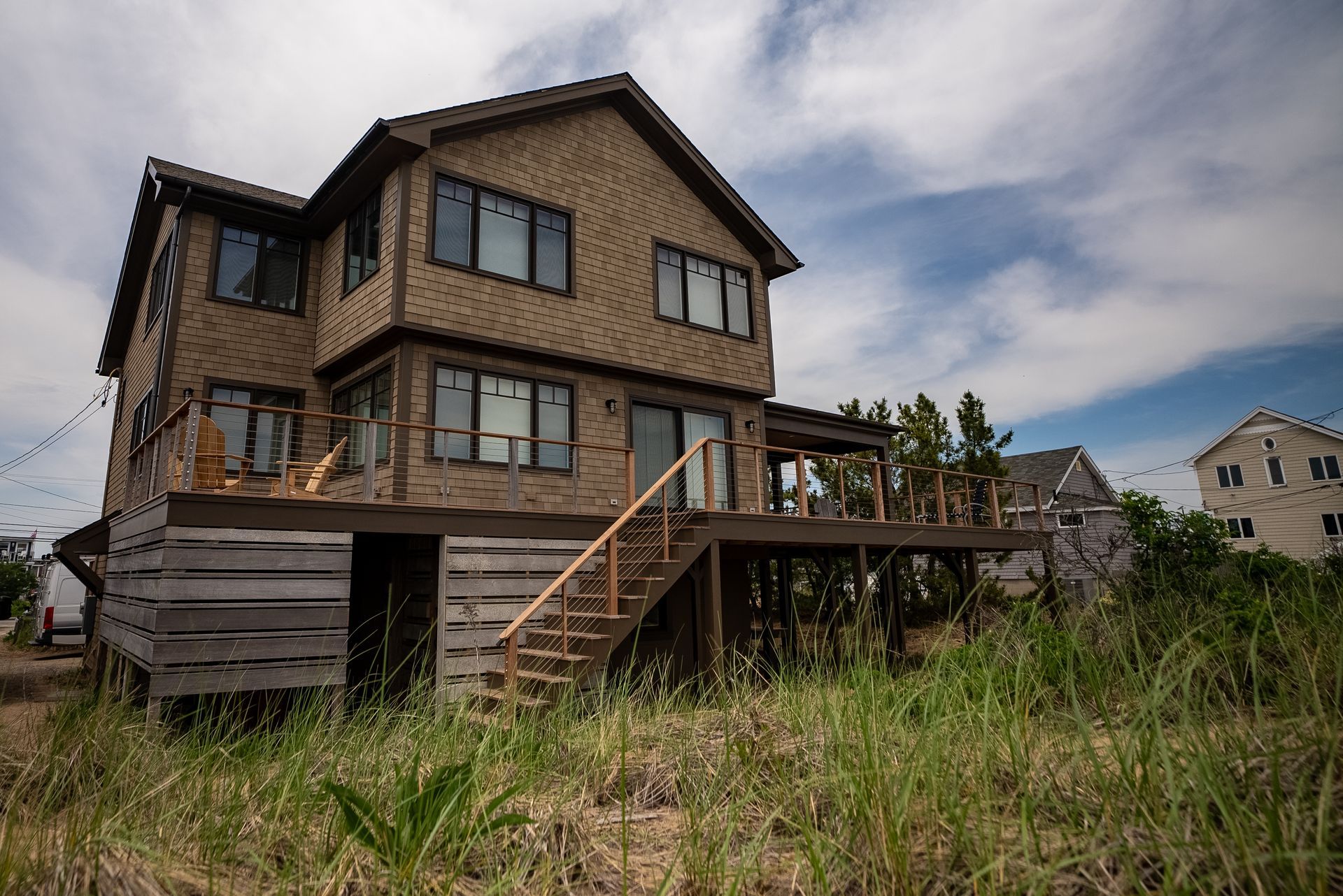 Modern two-story house with brown siding, deck, and stairs in an overgrown grassy lot under a cloudy sky