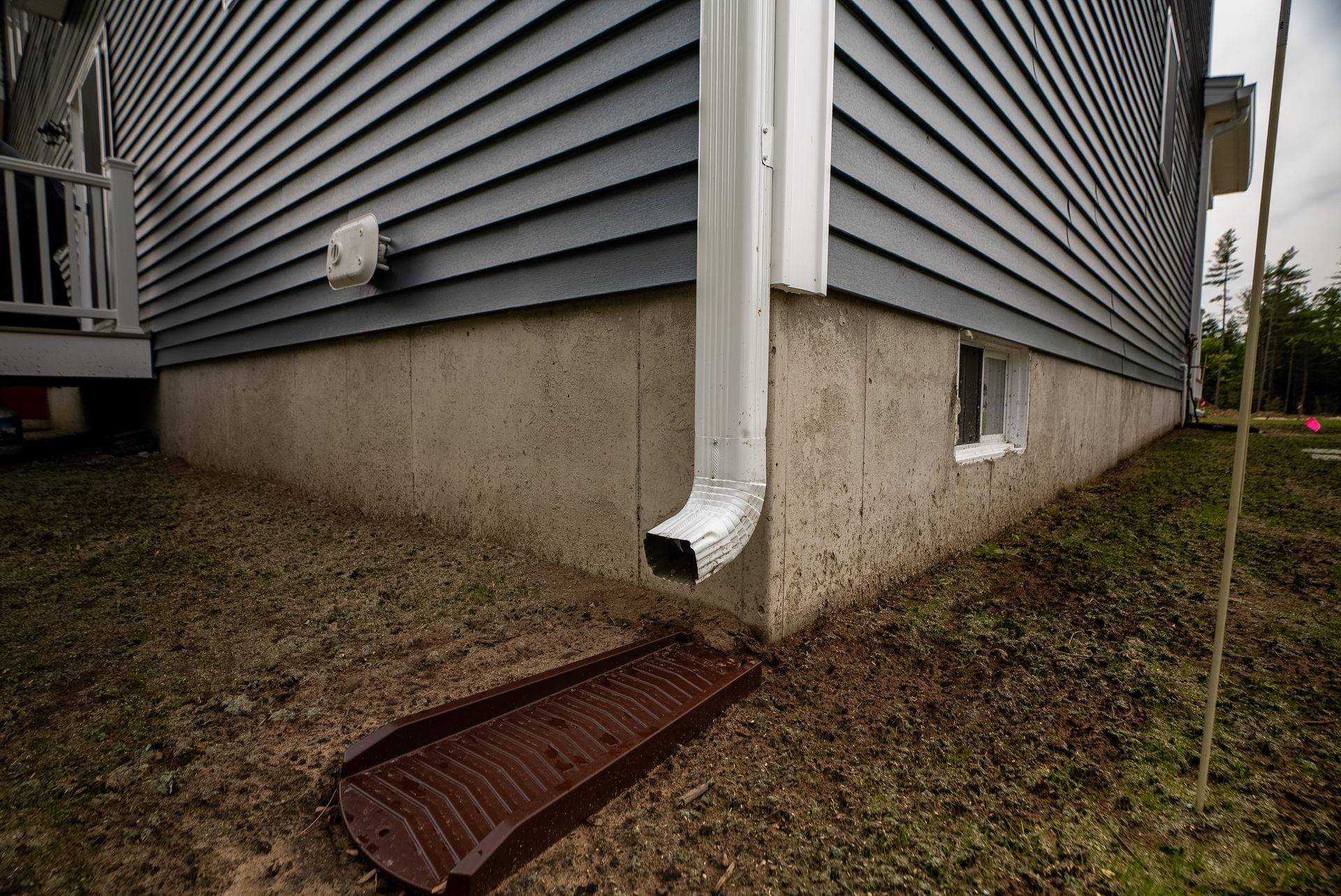 House corner with white downspout, concrete foundation, and rusted drain grate on damp grass