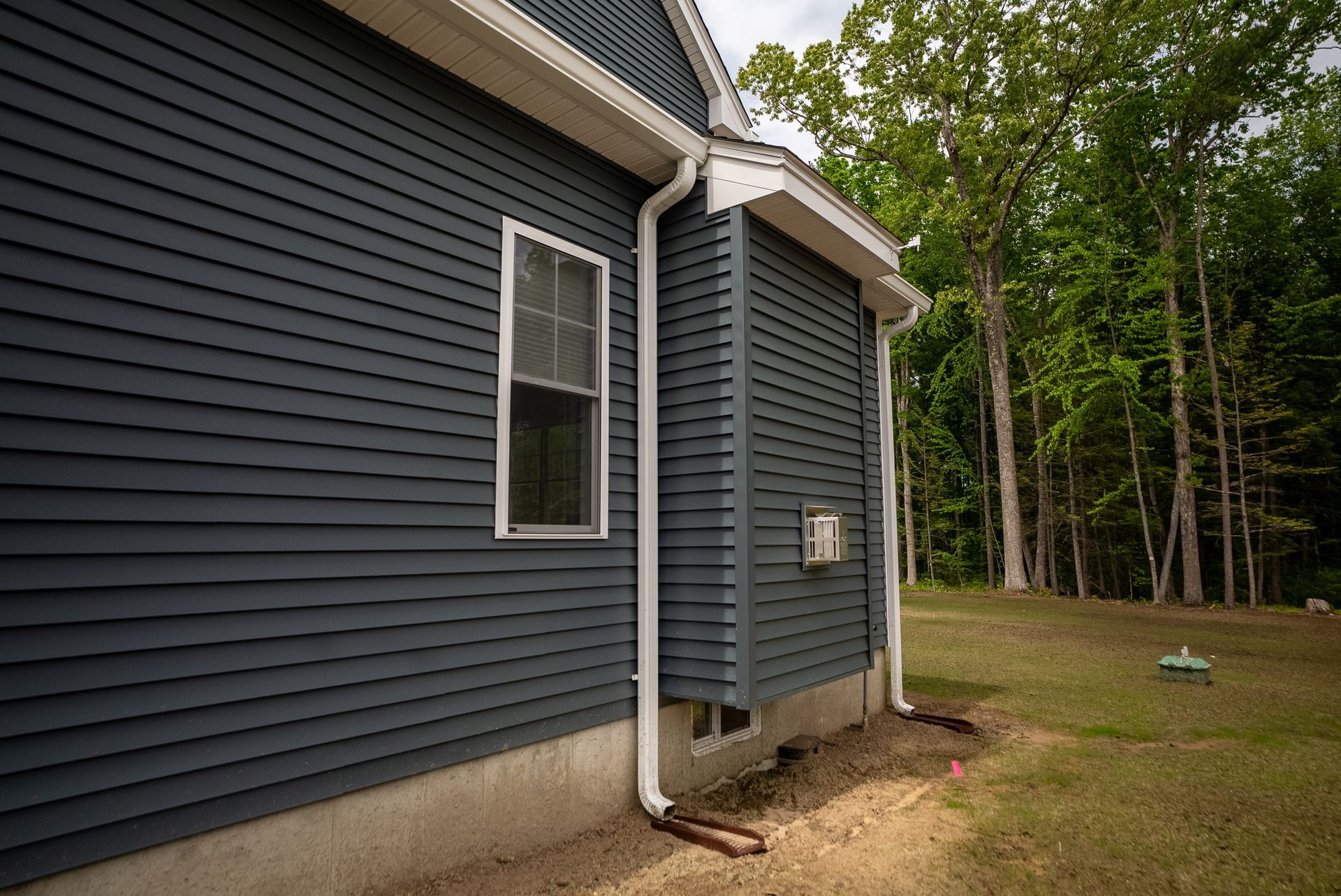 Side of a dark blue house with white trim and a downspout beside a grassy yard and trees