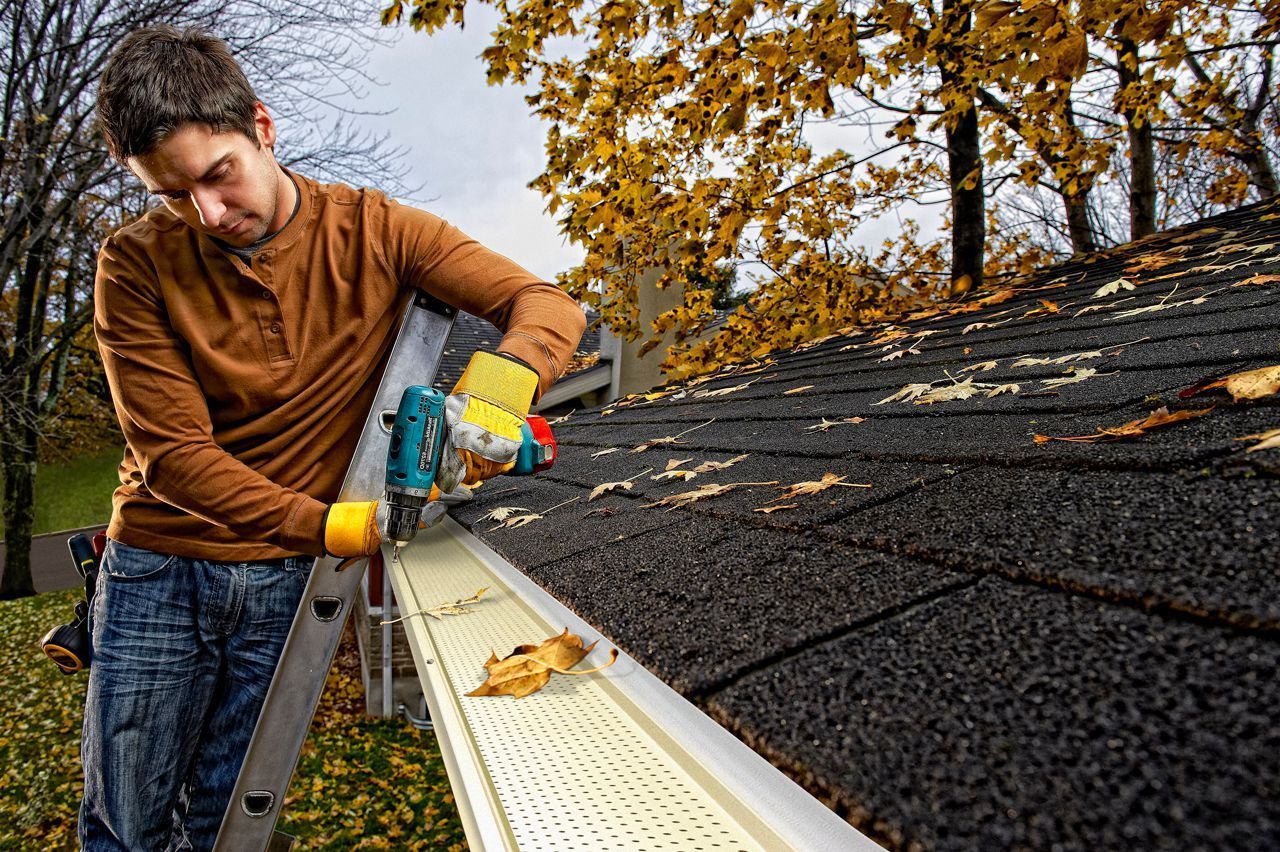 Man cleaning a roof gutter with a power tool, wearing gloves, leaves in gutter and trees in background.