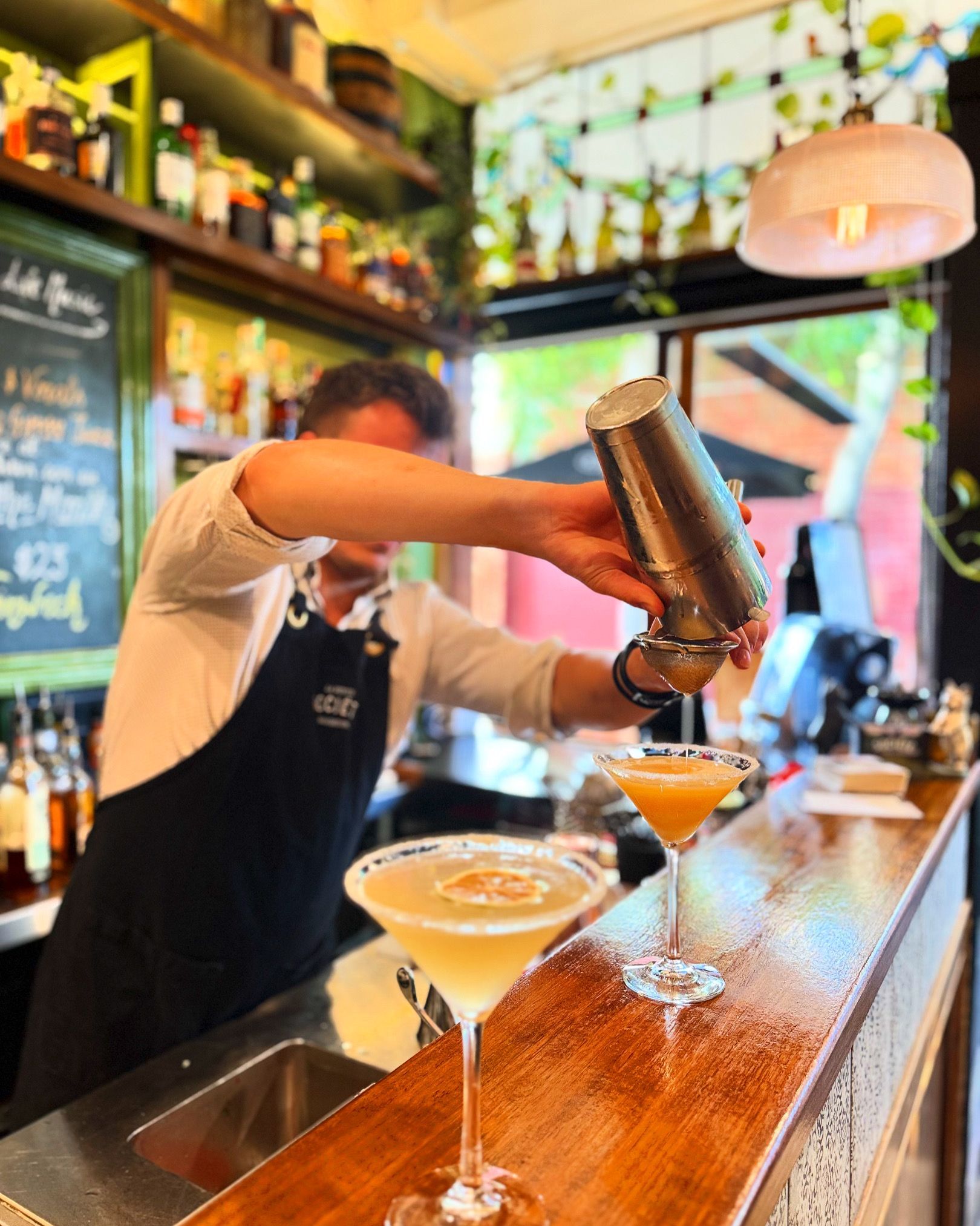 Bartender pouring a margarita into a cocktail glass on wooden bar. Bar setting, cocktails, man in apron.