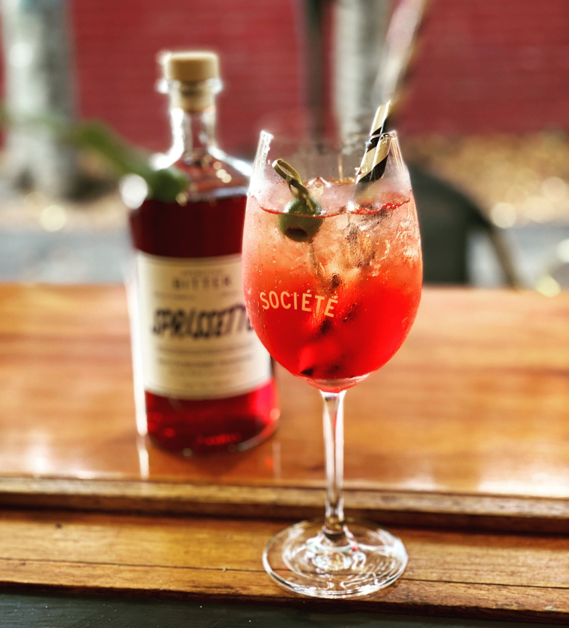Red spritz cocktail in a stemmed glass with a bottle of Spritzetto in the background on a wooden bar.
