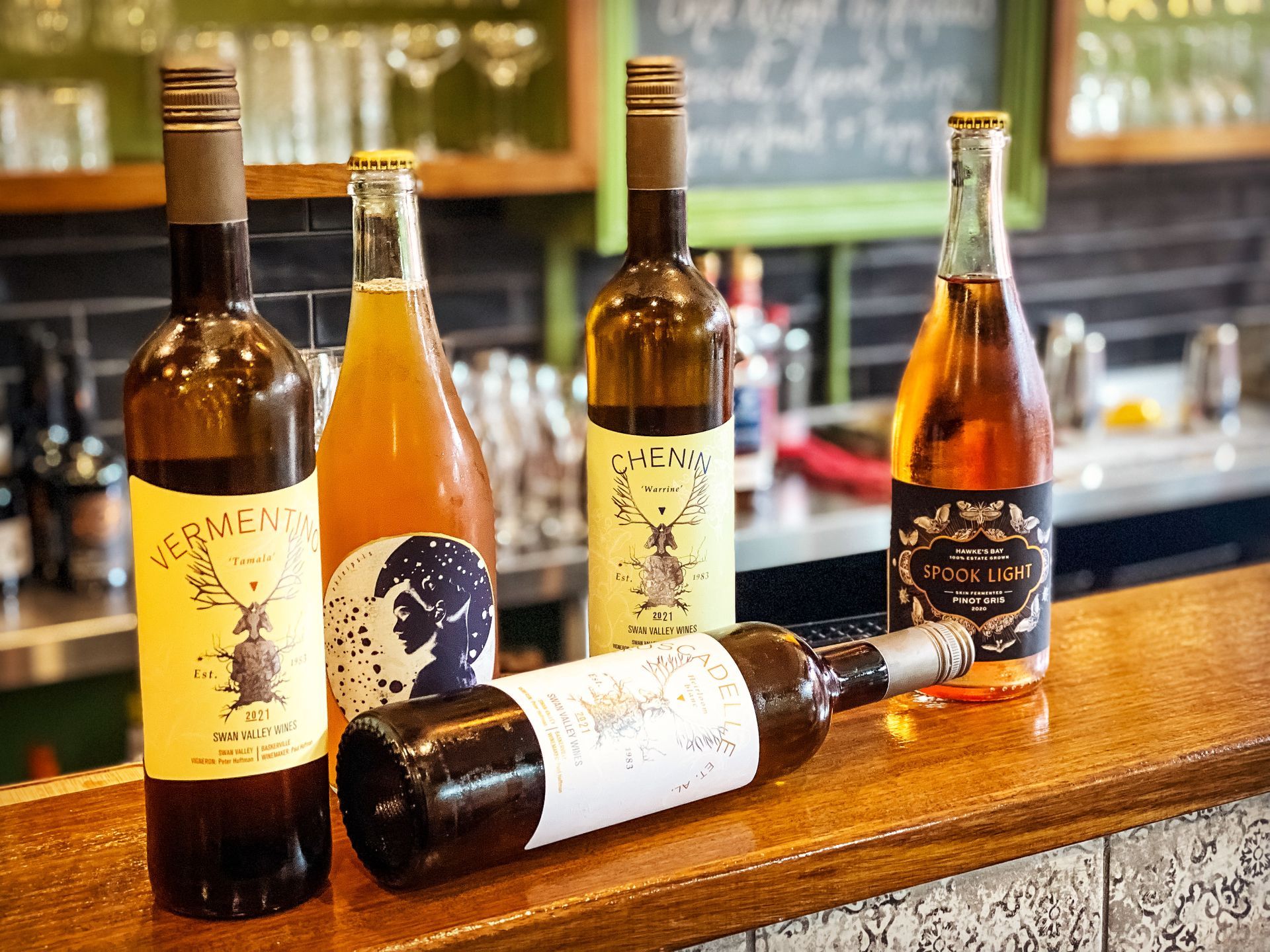 Bottles of wine on a bar. Various labels and colors, some standing, one lying down. Blurred green background.
