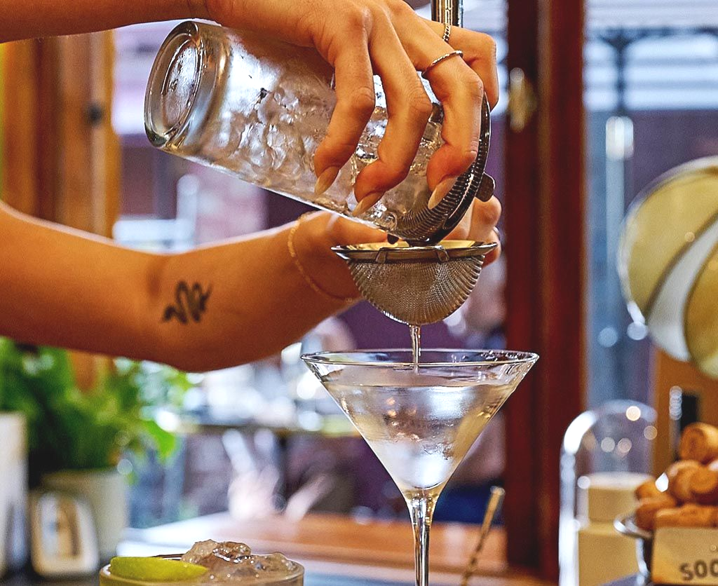 Bartender strains cocktail into a martini glass, bar setting.