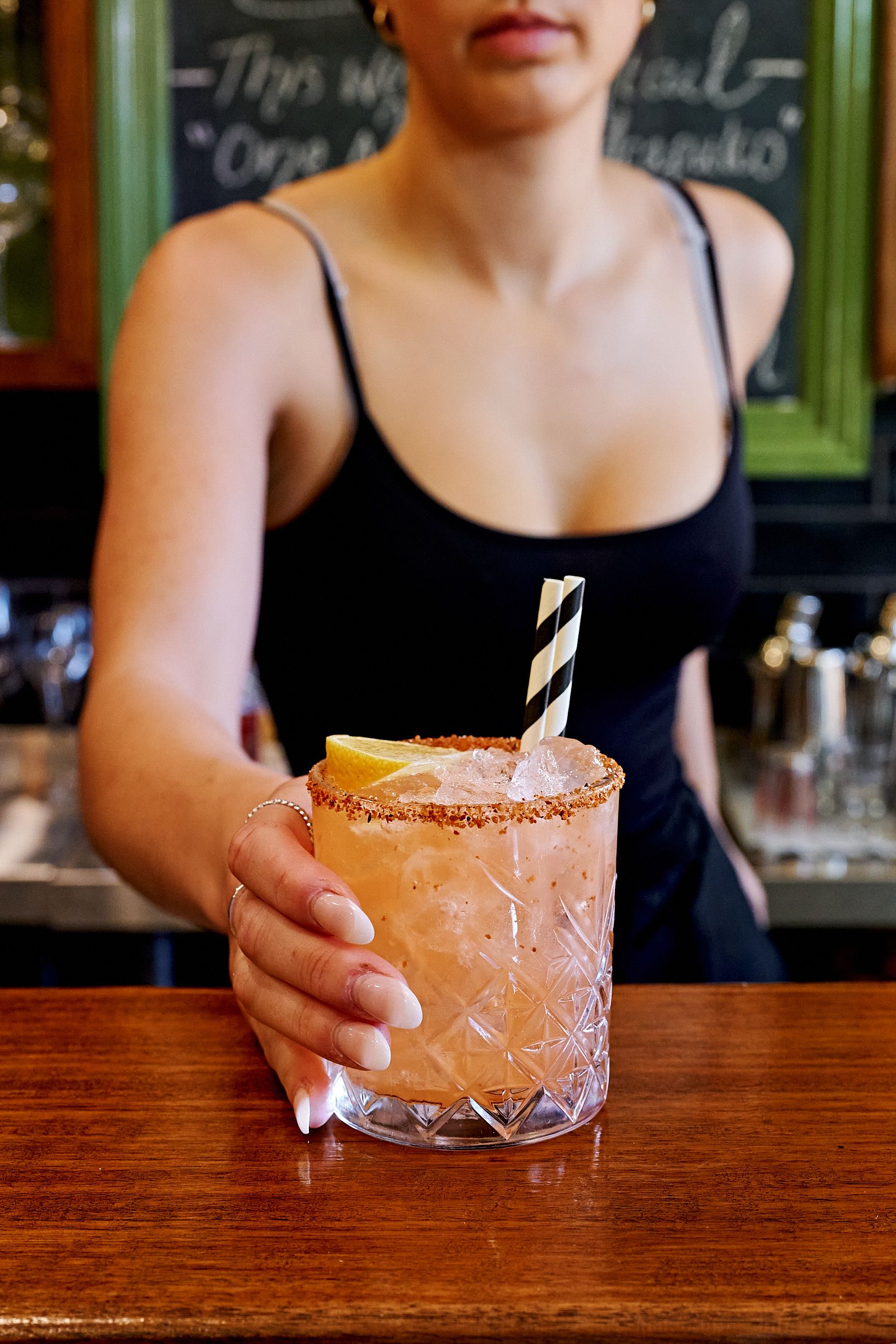 Bartender in black top serving a cocktail with a salted rim and straw over a wooden bar.