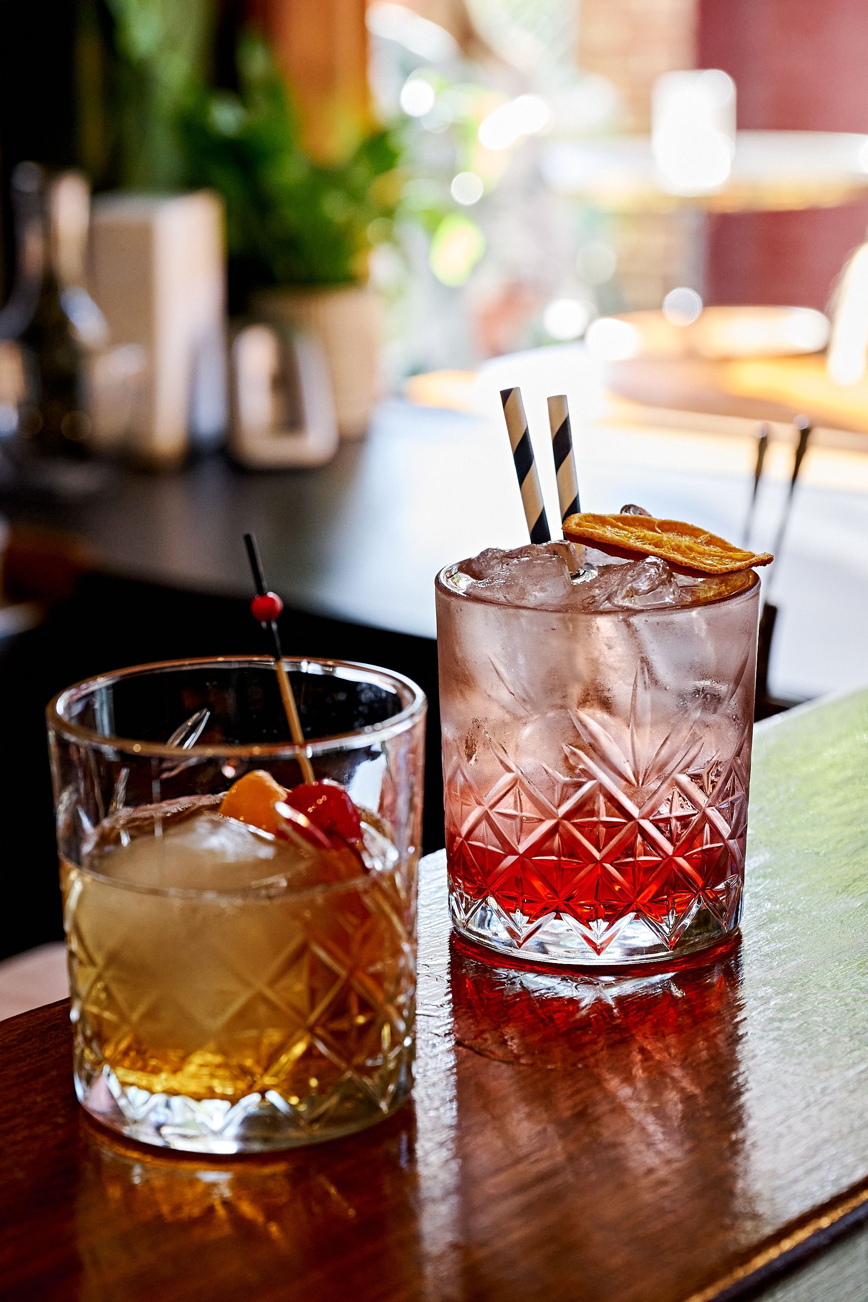 Two cocktails on a bar, one amber, one pink, both with ice.