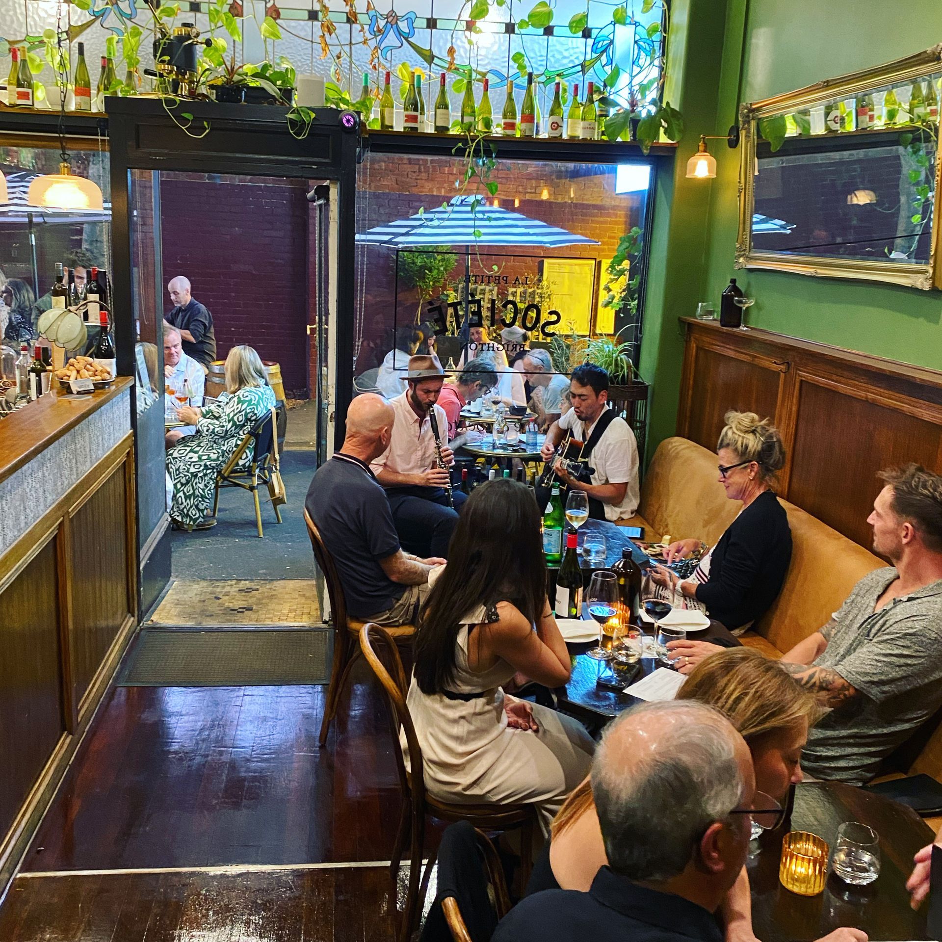 People dining and drinking at a wine bar, some outdoors, with green walls and floral decorations and a jazz band playing.
