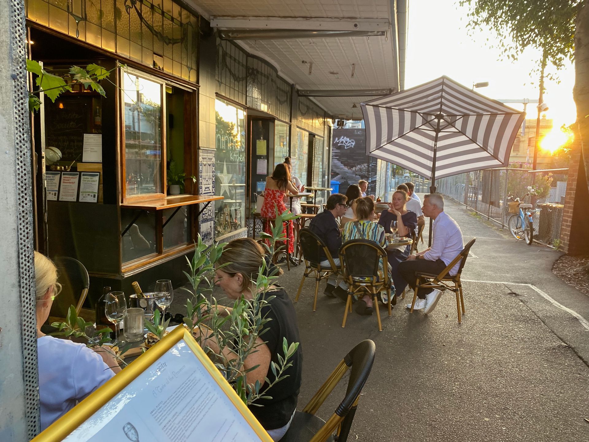 Outdoor restaurant with people seated at tables, eating and talking. Striped umbrella provides shade; late afternoon sun.
