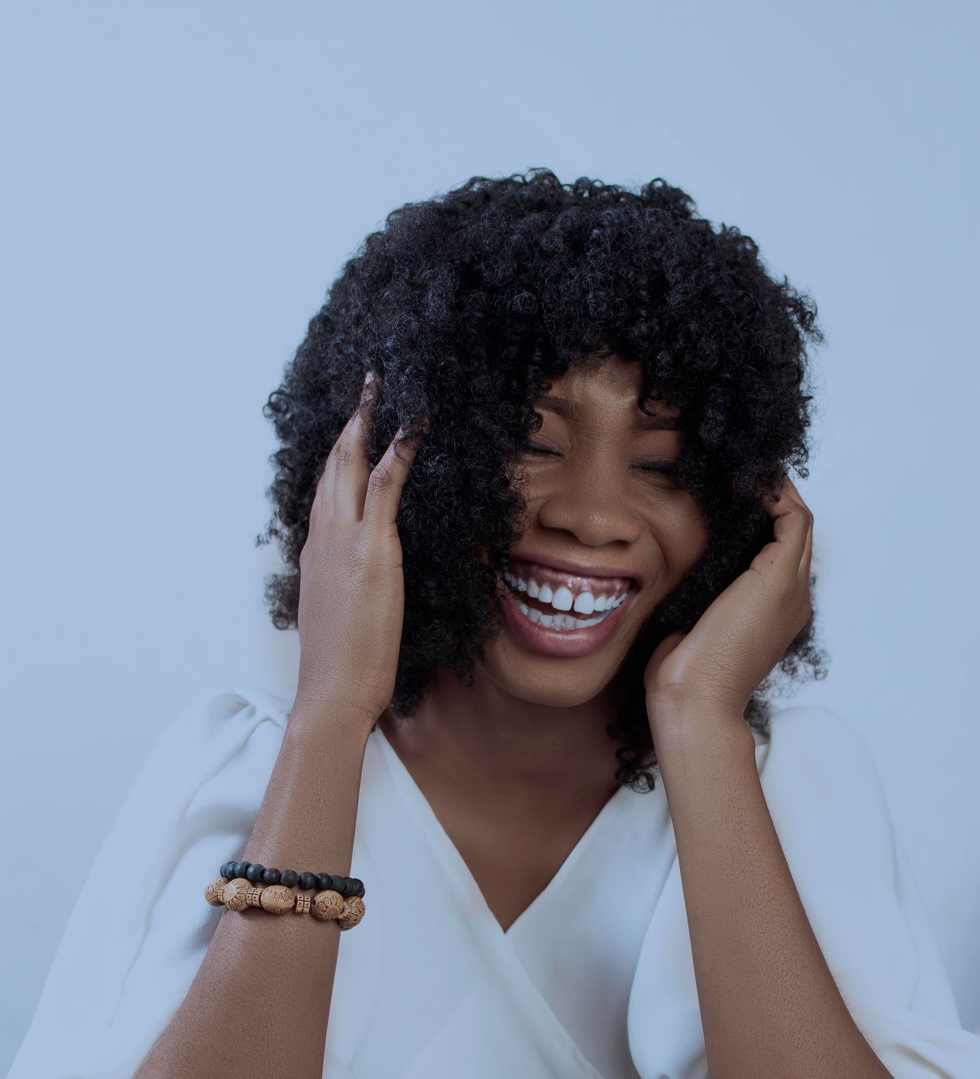 A woman with curly hair is smiling and covering her ears with her hands.