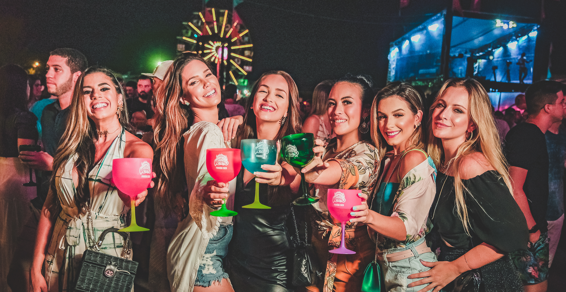 Um grupo de mulheres estão de pé, uma ao lado da outra, segurando taças de vinho em uma festa.