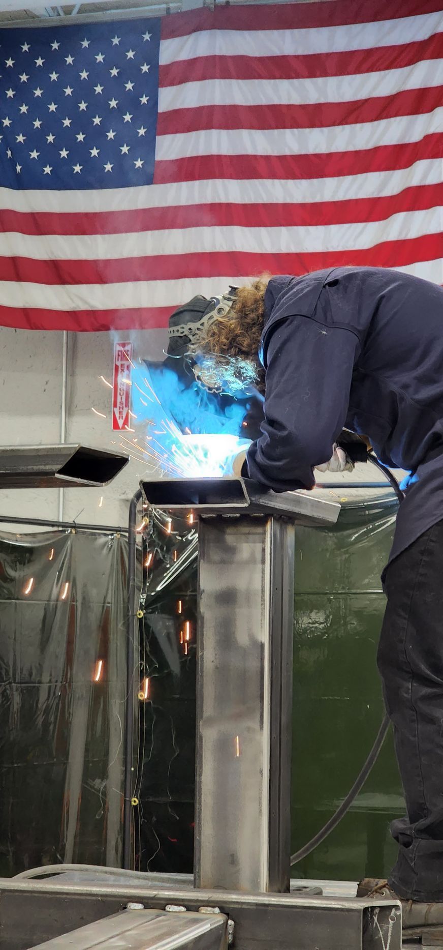 A man is welding a piece of metal in front of an american flag.