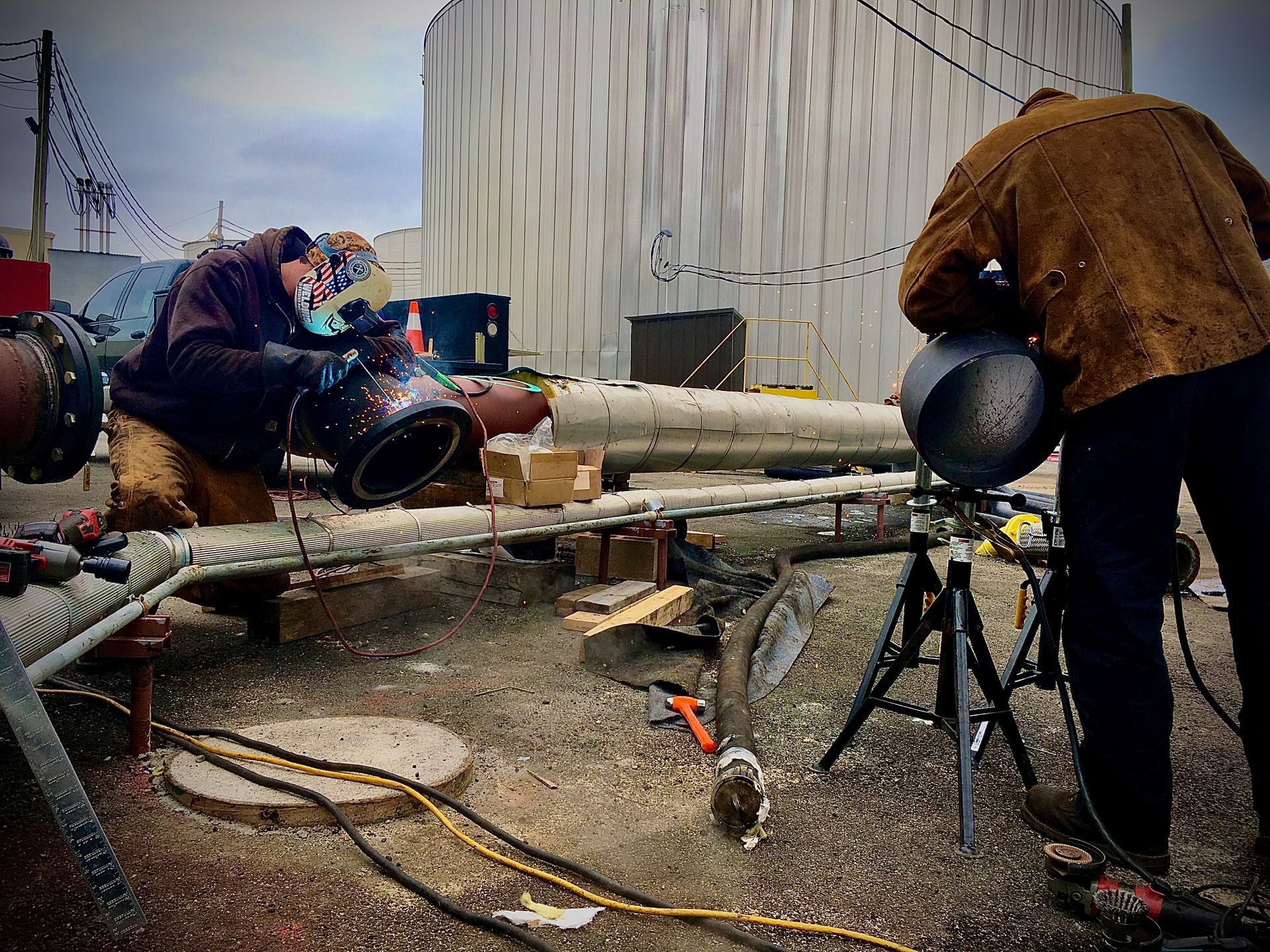 Two men are welding a pipe in a yard.