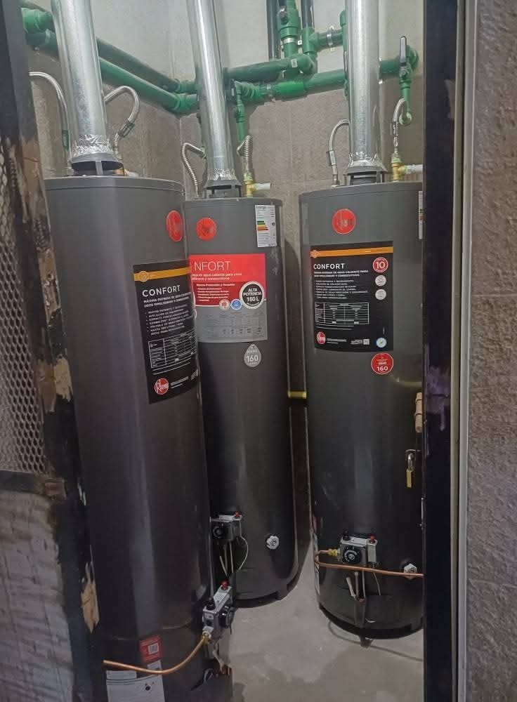 Tres calentadores de agua grises en un cuarto de servicio con tuberías de ventilación plateadas y tuberías verdes.