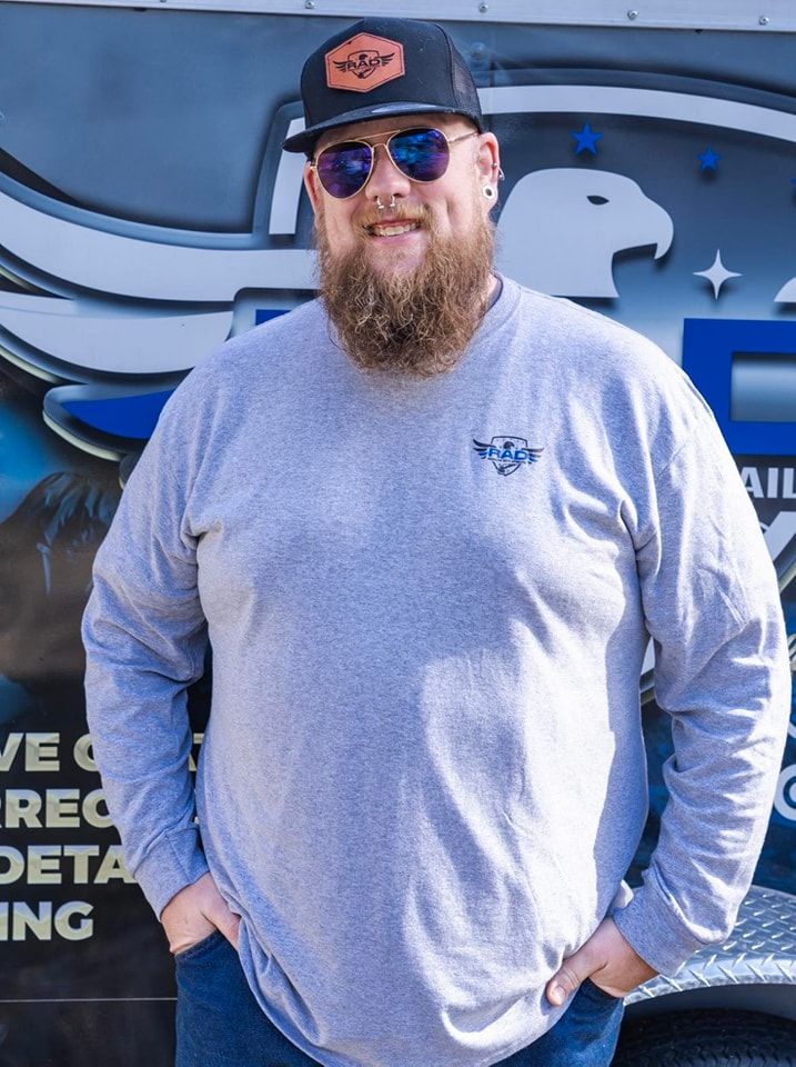 A man with a beard wearing sunglasses and a hat is standing in front of a truck.