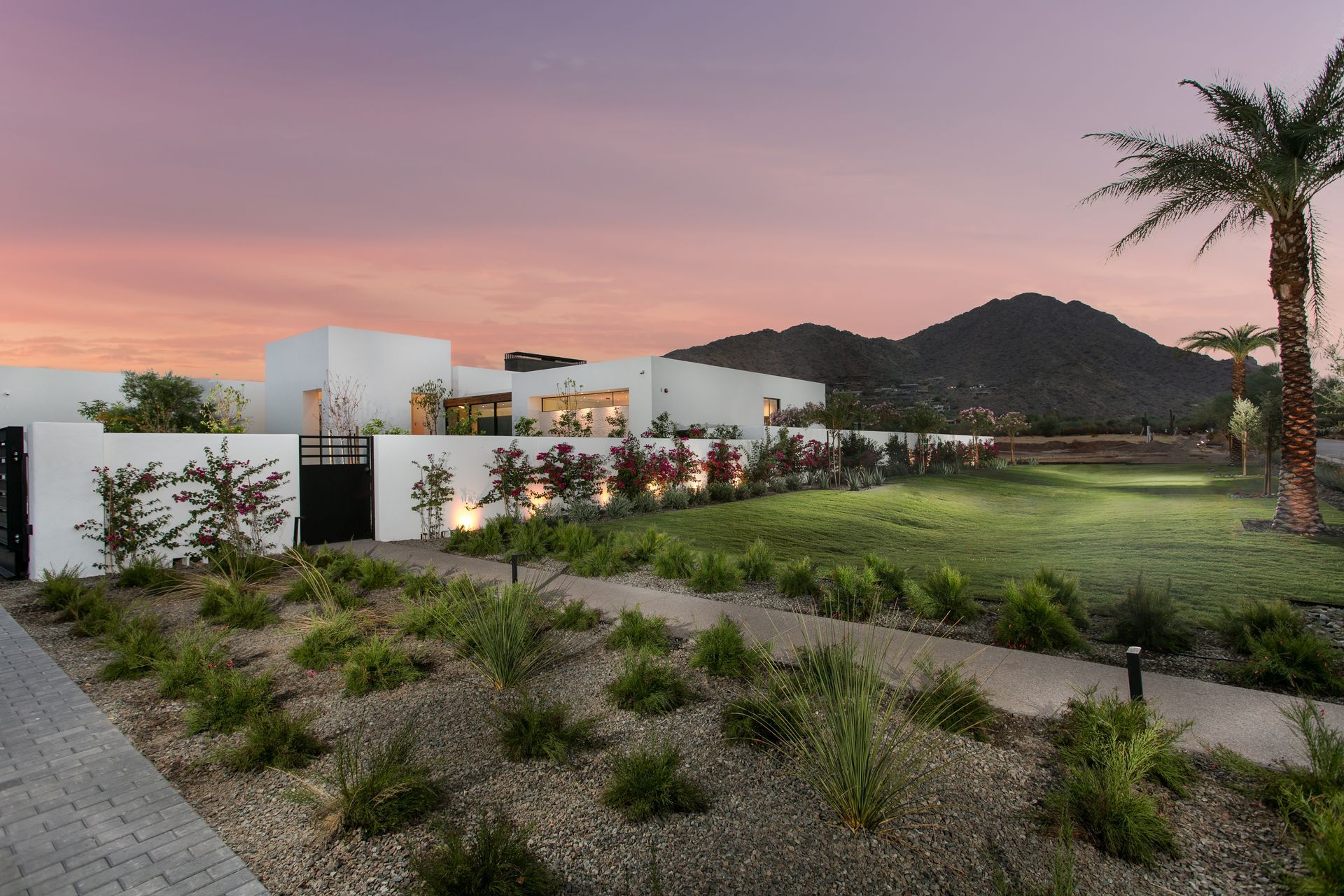 White house with a palm tree, green lawn, and mountain backdrop at sunset.