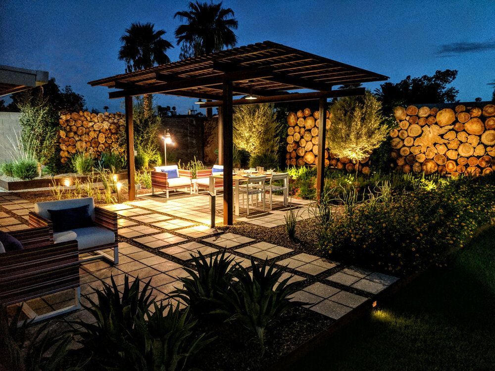 Night-lit patio with pergola, table, chairs, and stacked log wall. Landscaping with soft illumination.