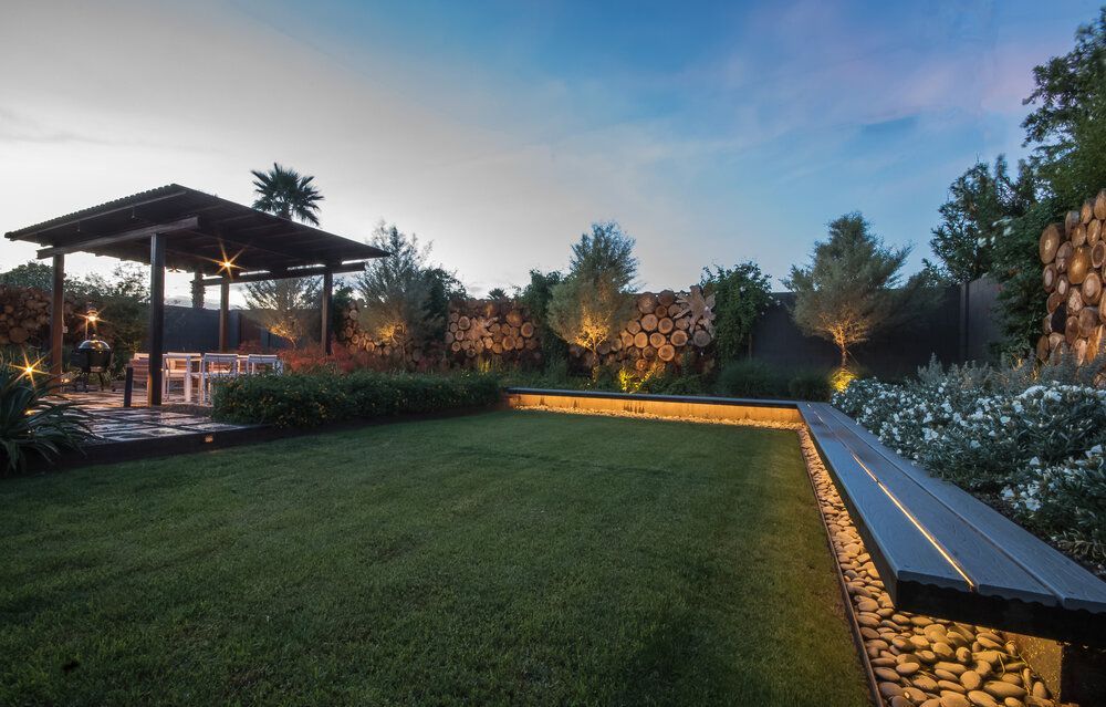 A well-lit backyard at dusk with a gazebo, bench, and manicured lawn, with trees along the border.