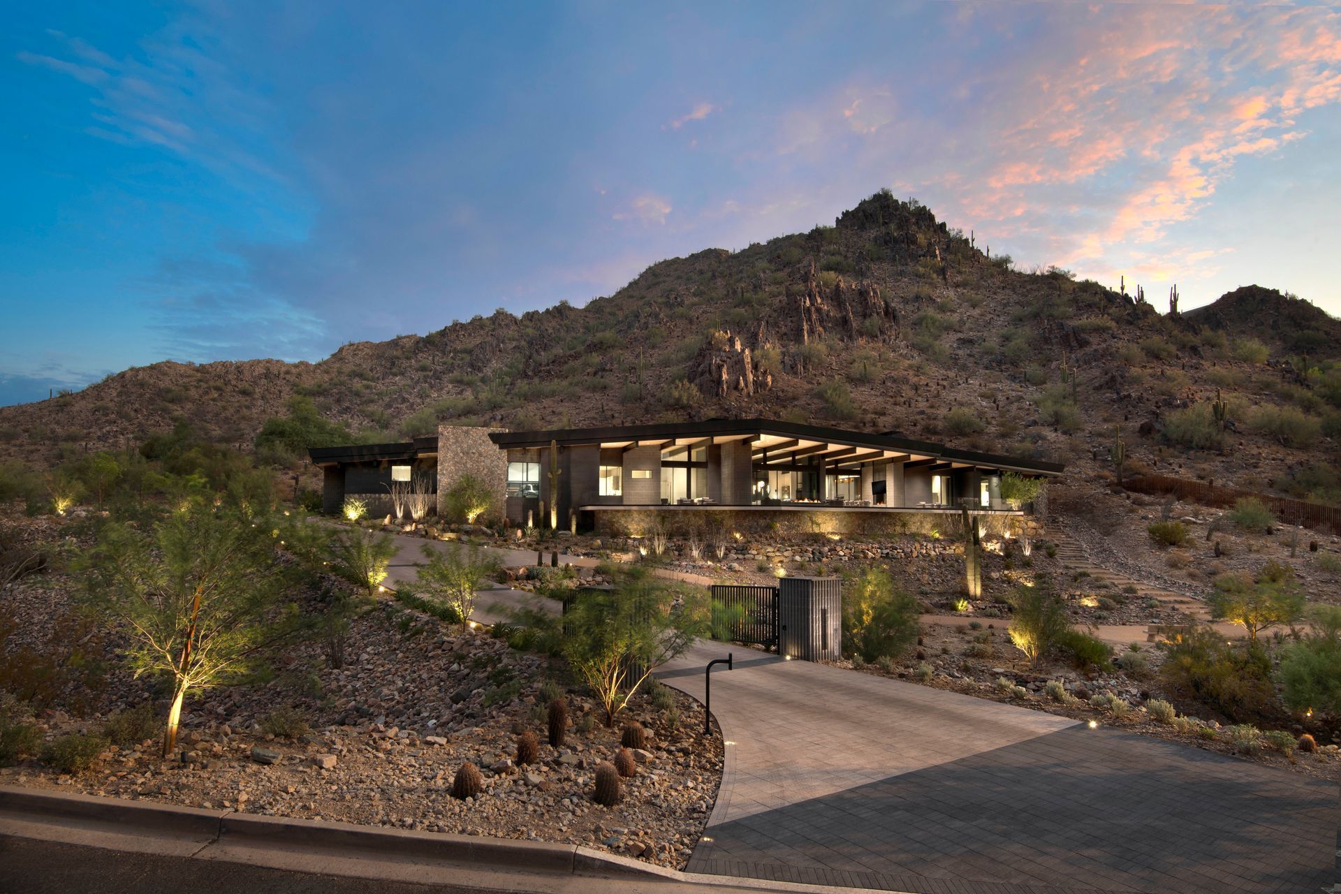Modern home nestled at the base of a mountain, illuminated at dusk. Driveway leads to the entrance.