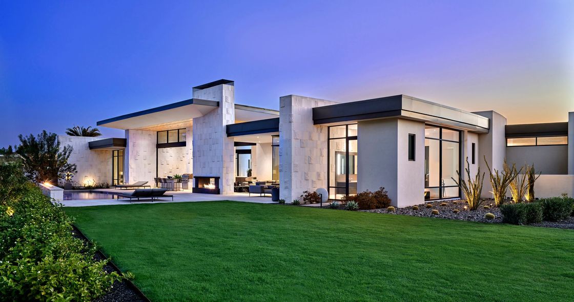 Modern, white stucco house with a green lawn, pool, and fireplace at dusk.