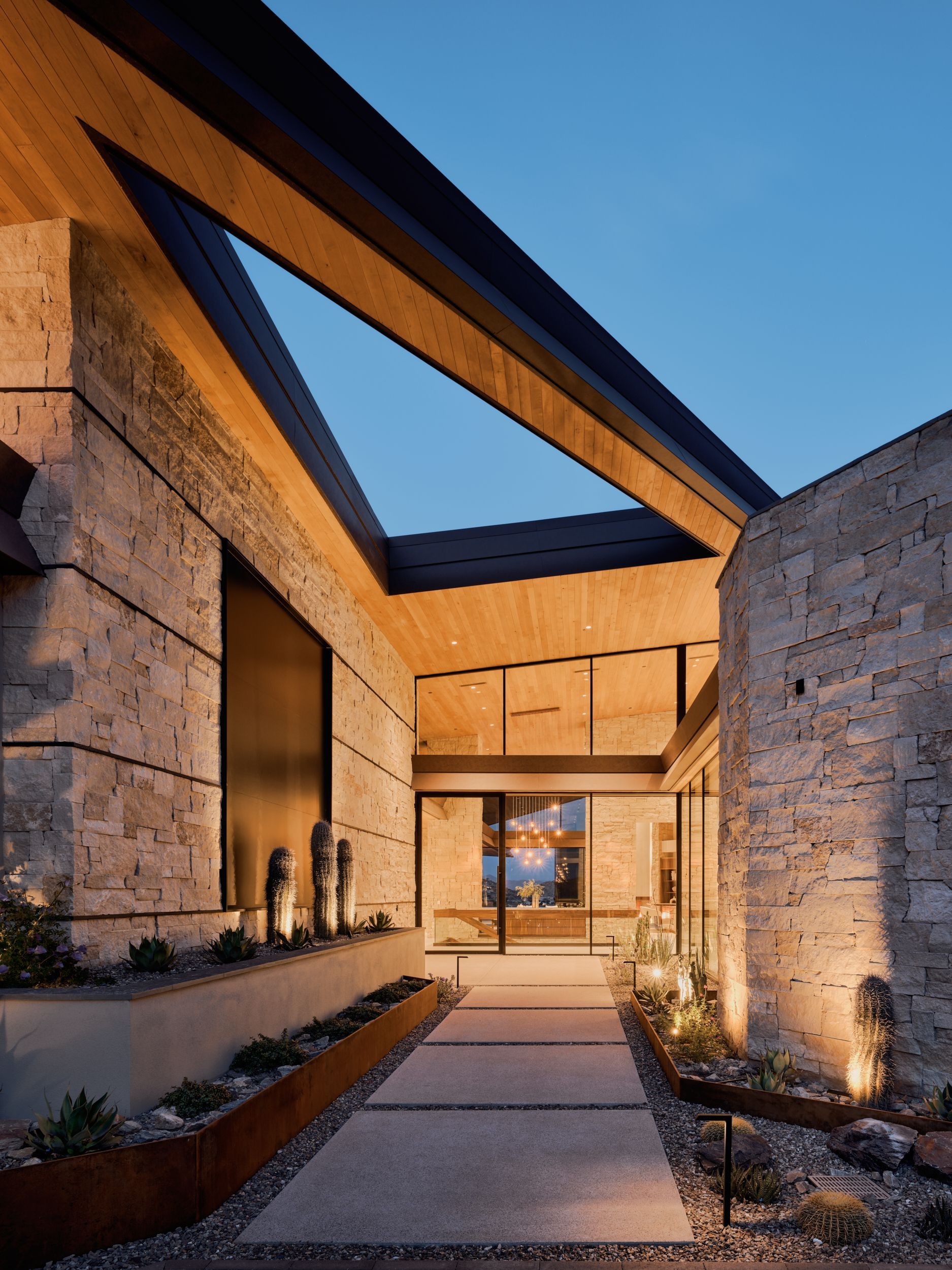 Stone-walled entrance to a modern home with a dark sloped roof and pathway illuminated by warm lights.
