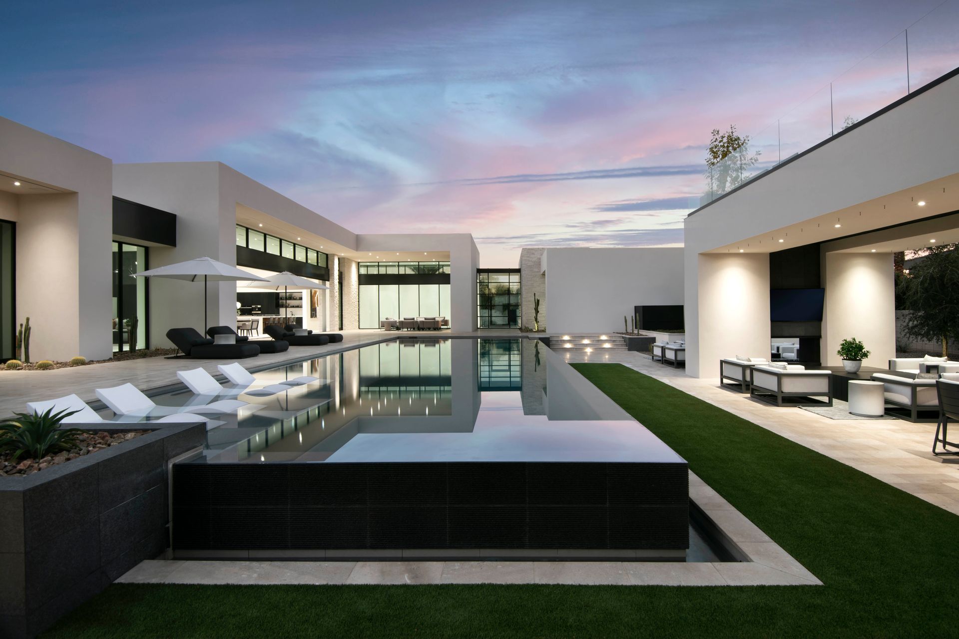 Modern luxury home with pool at dusk; white exterior, lawn, outdoor seating, and colorful sky.