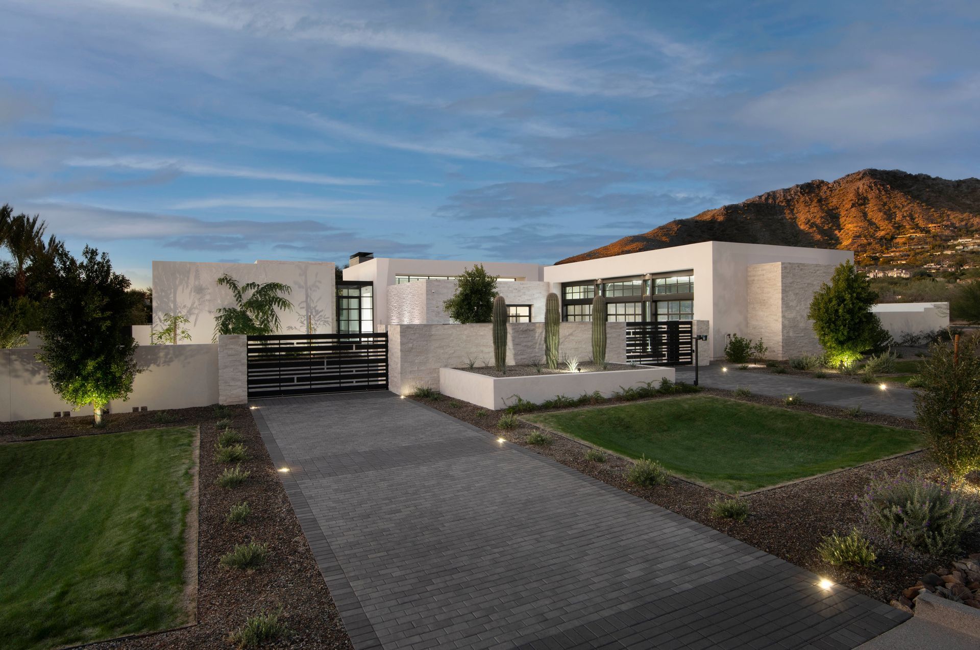Modern white house with dark gray driveway and landscaping, mountain backdrop at dusk.