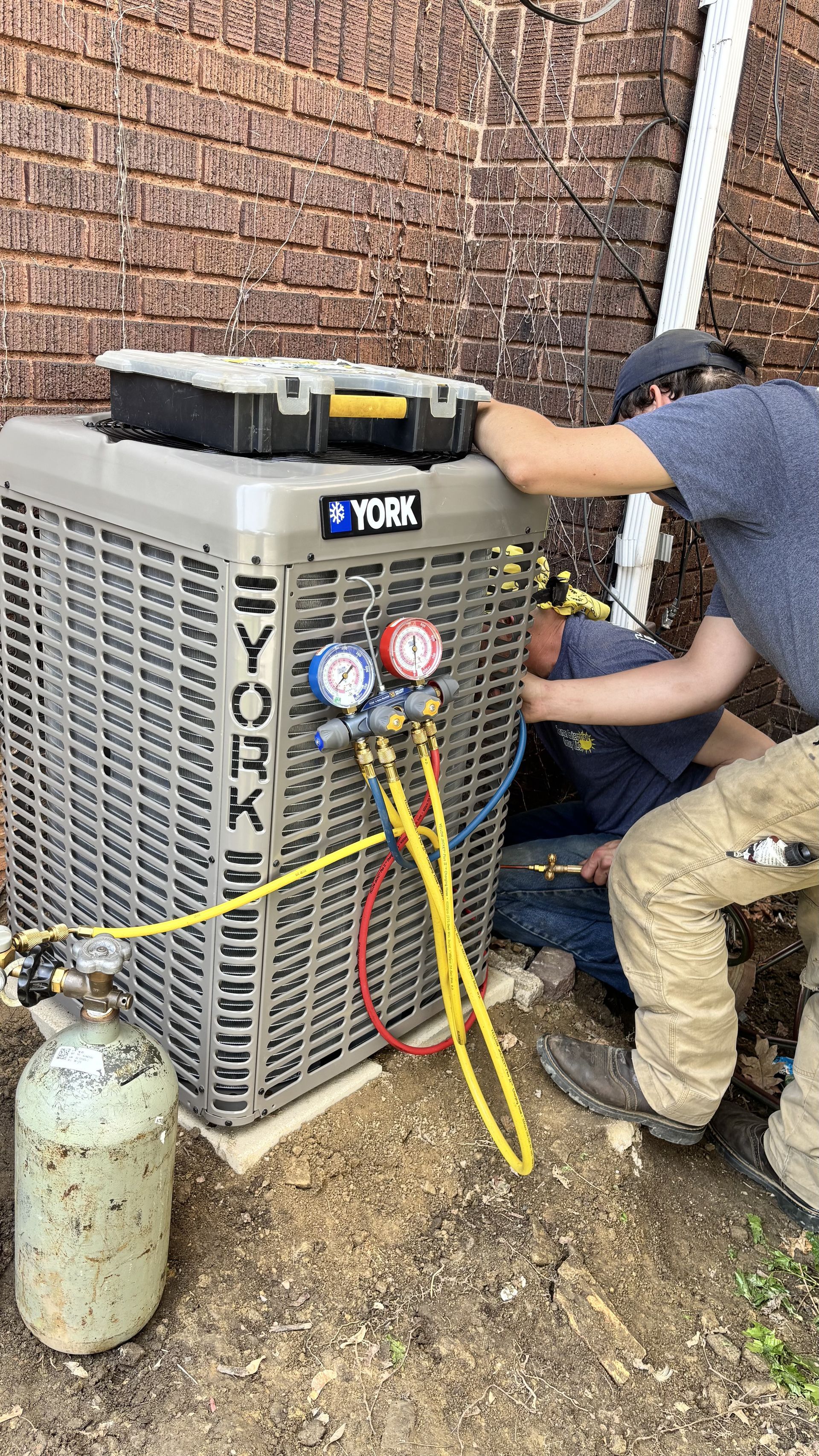Two men are working on an air conditioner outside of a brick building.