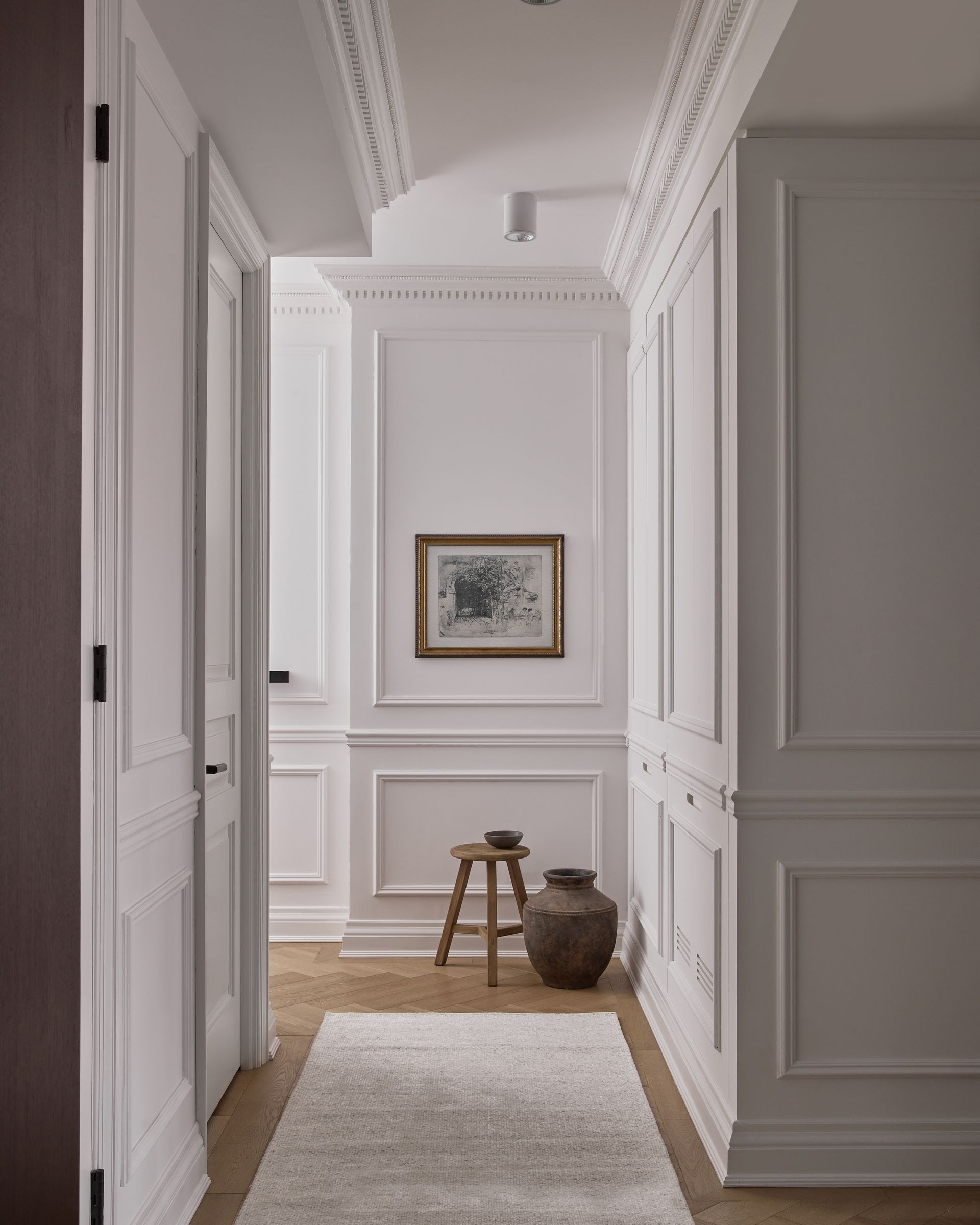 A hallway with white walls , a rug , a vase , a stool and a picture on the wall.