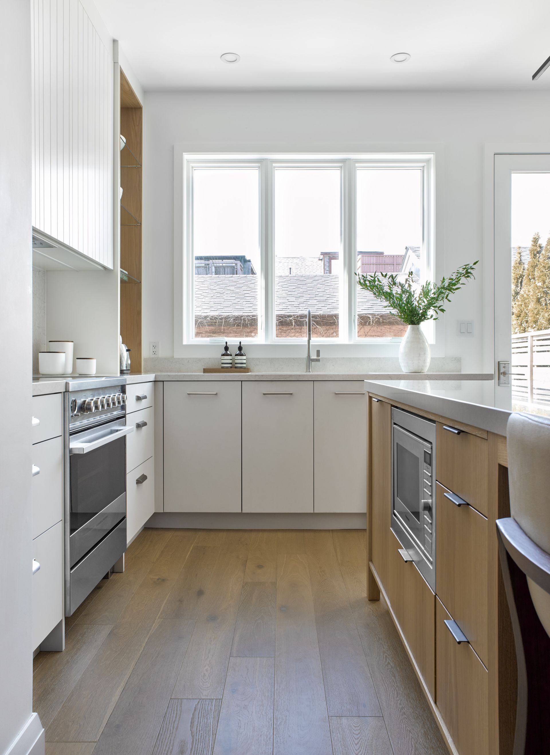 A kitchen with white cabinets , wooden floors , stainless steel appliances and a large window.
