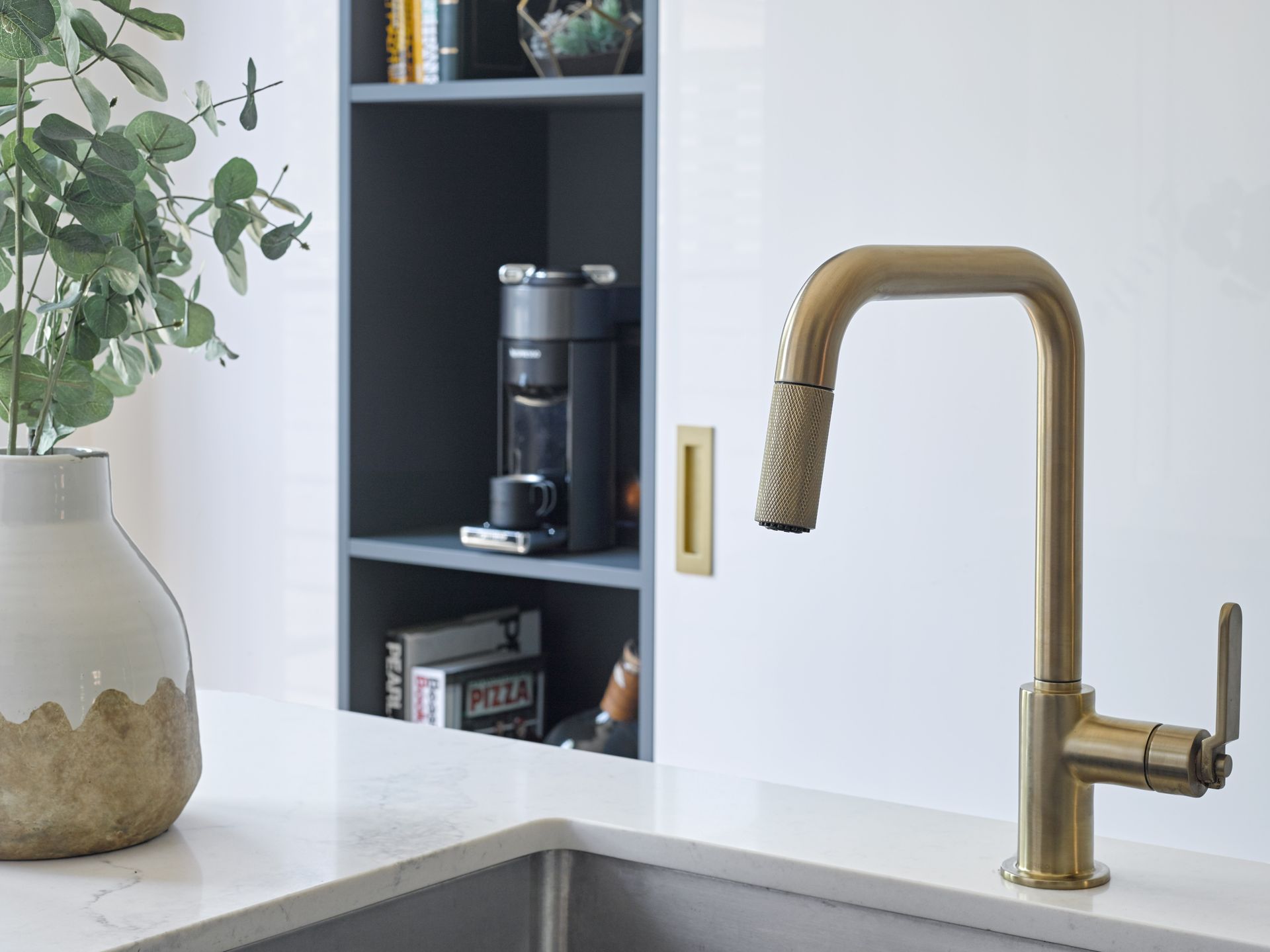 A kitchen sink with a brass faucet and a coffee maker on a shelf.