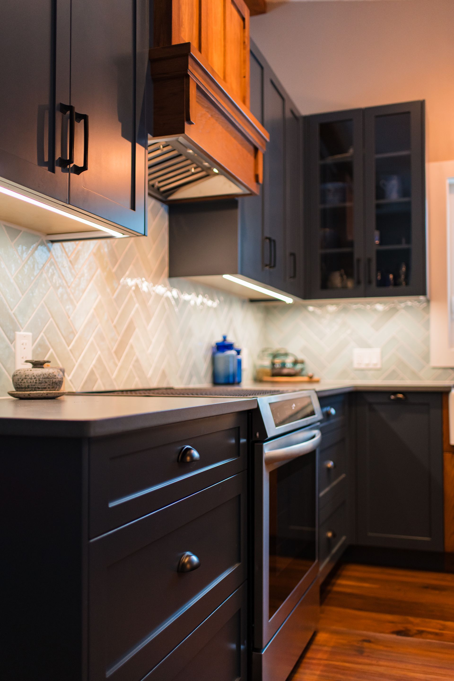 Modern kitchen with dark cabinets, stainless oven, white backsplash, and warm wood flooring.