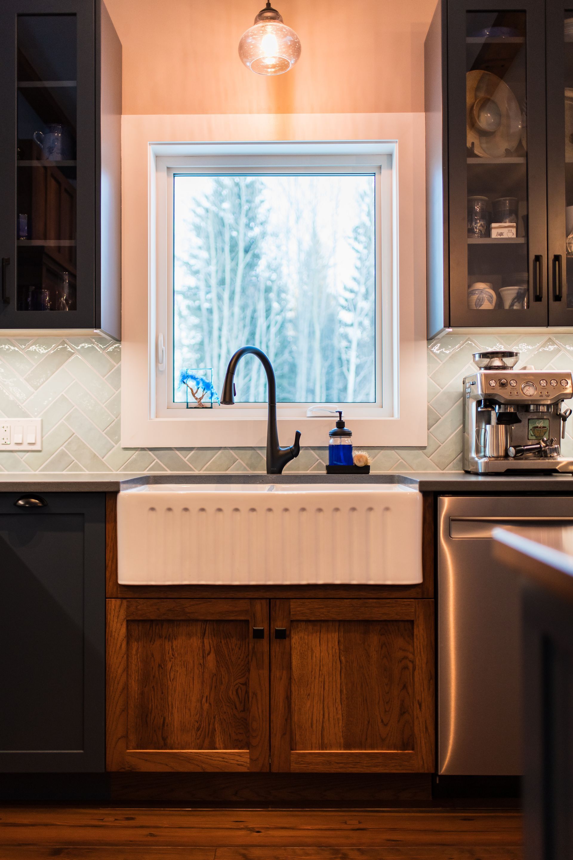 Modern kitchen sink beneath a window, with dark cabinets, white backsplash, and wood-paneled base cabinets.