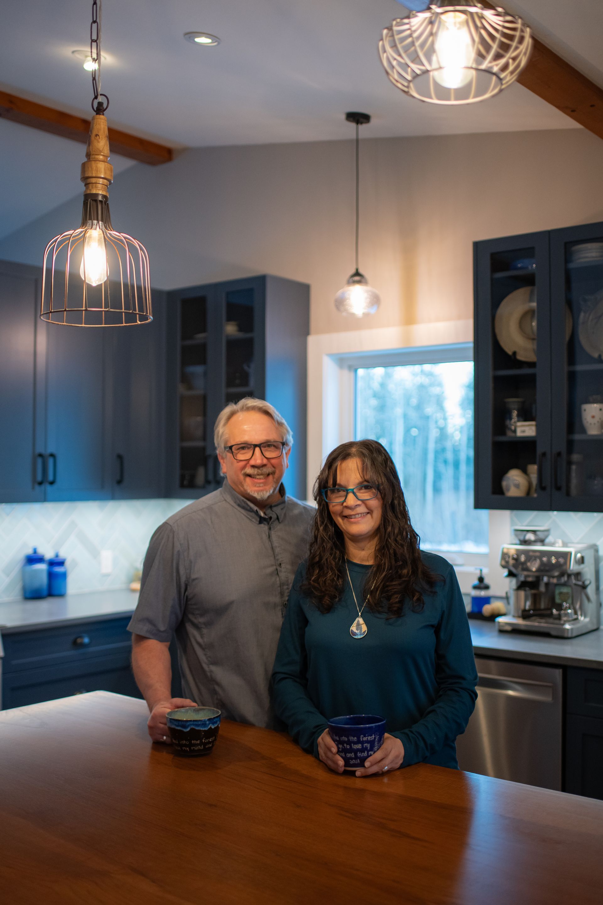 Two people standing in a modern kitchen beside a wooden table, with pendant lights and blue cabinets.
