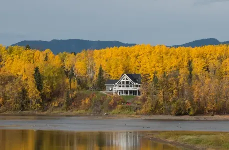 Lakeside house in front of vivid yellow autumn trees, with mountains and water reflections in the background