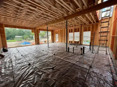 Interior of a framed house under construction with exposed beams, plastic-covered floor, and ladders.