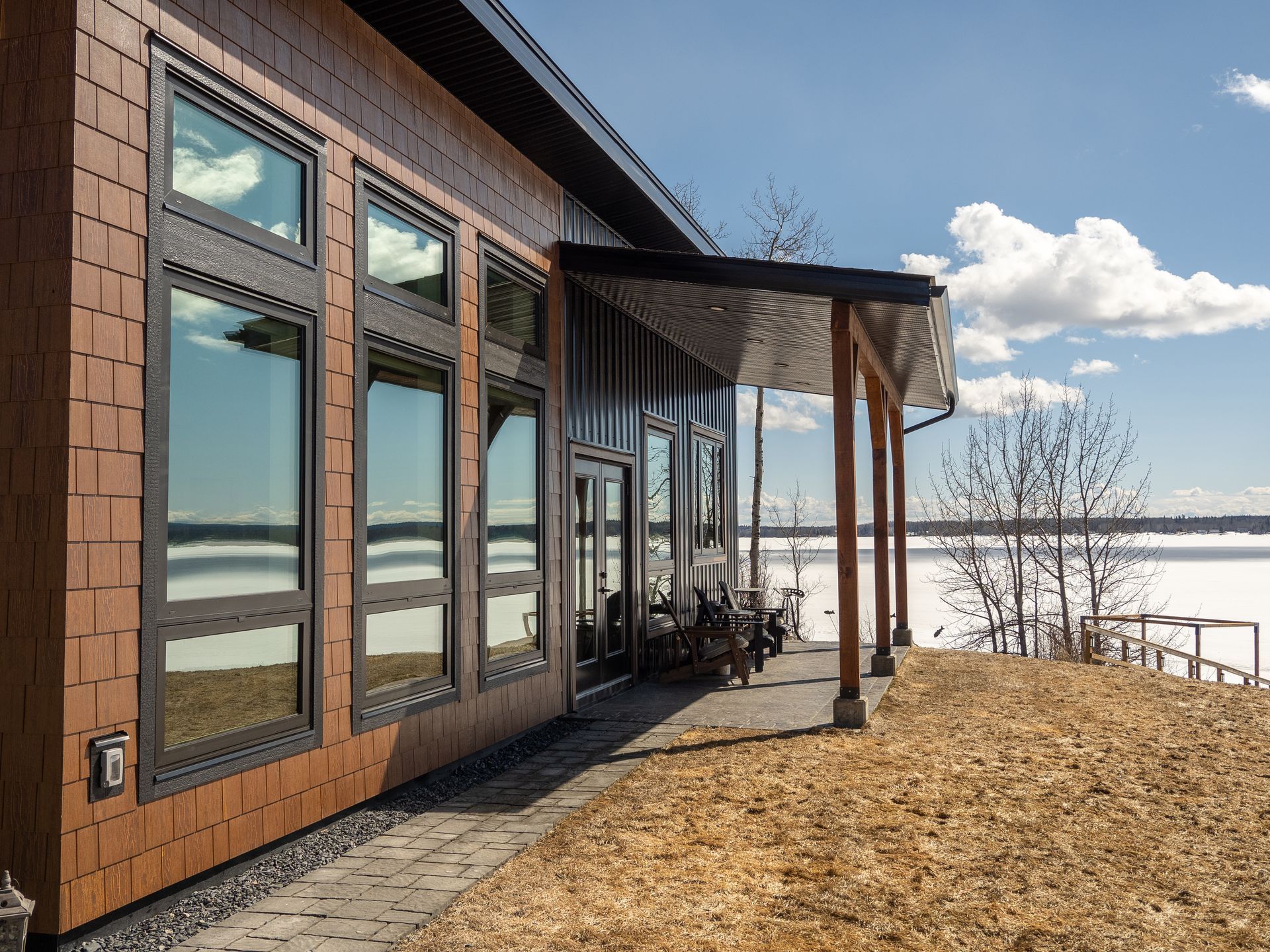 Modern lakeside house with large windows, wooden siding, and a covered porch on a snowy shoreline