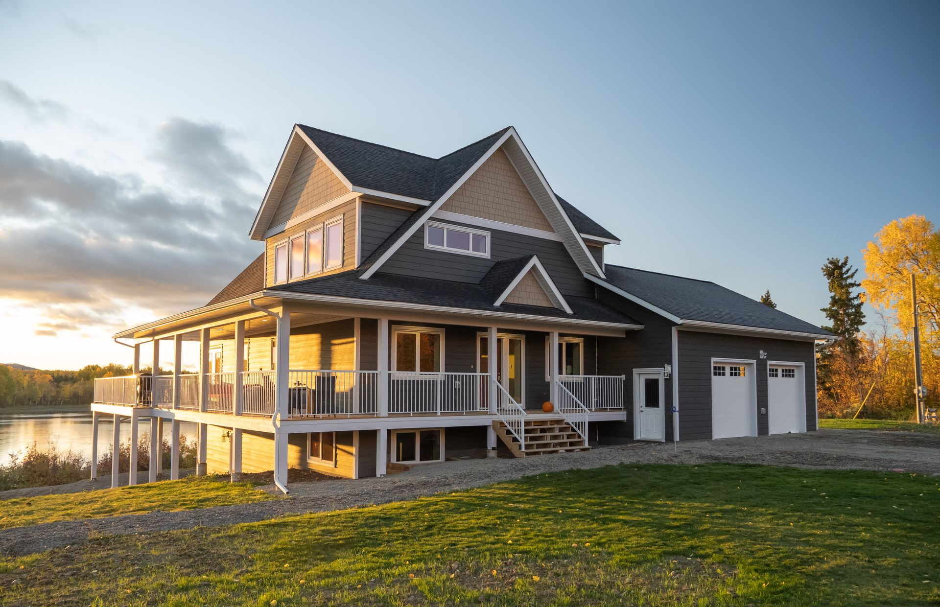 Modern farmhouse with wraparound porch at sunset, beside a grassy field and lake