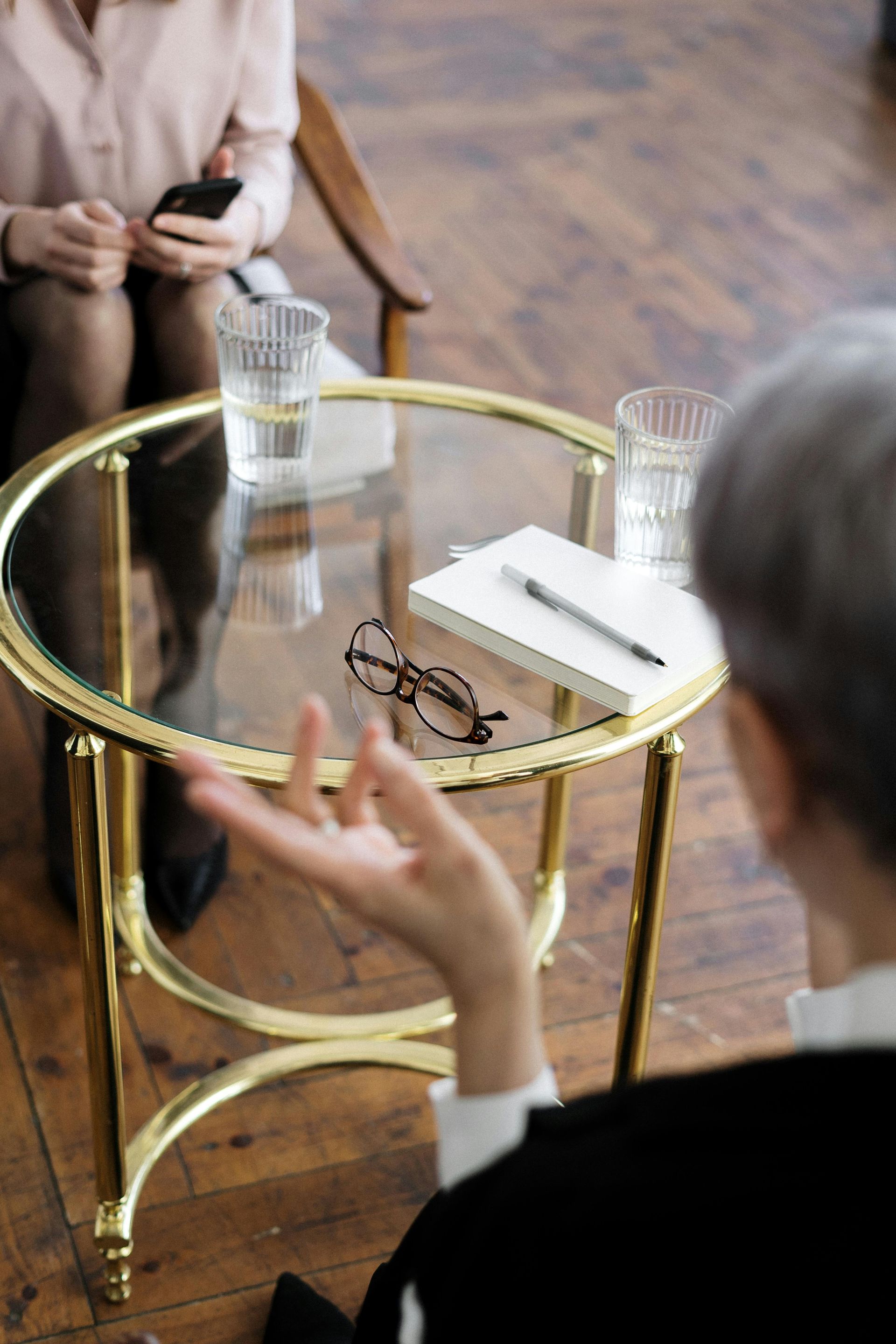 Two people seated at a small glass table with water glasses and a notebook in a wood-floored room