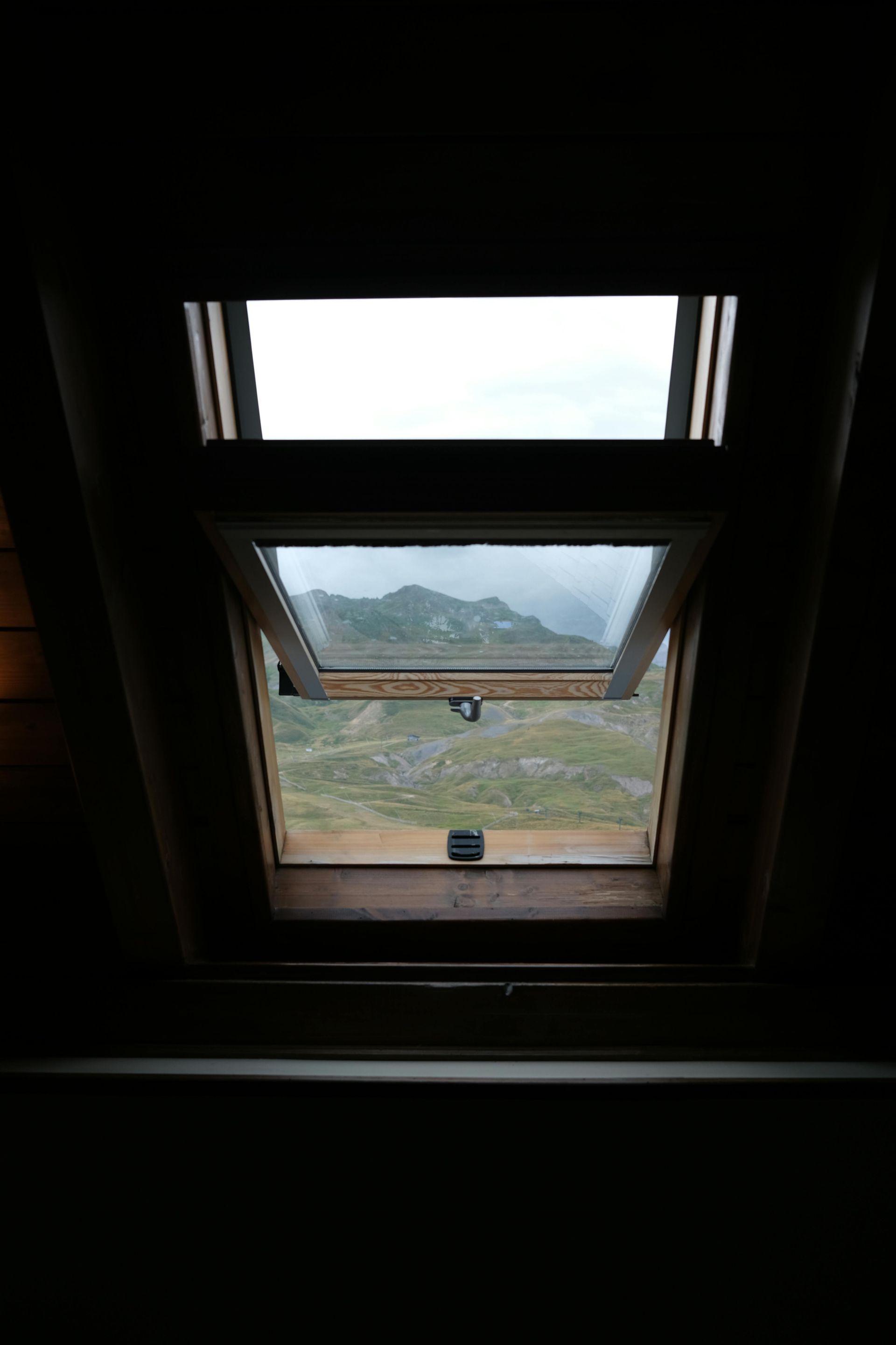 Dark interior view through a window to a mountain landscape under a cloudy sky.