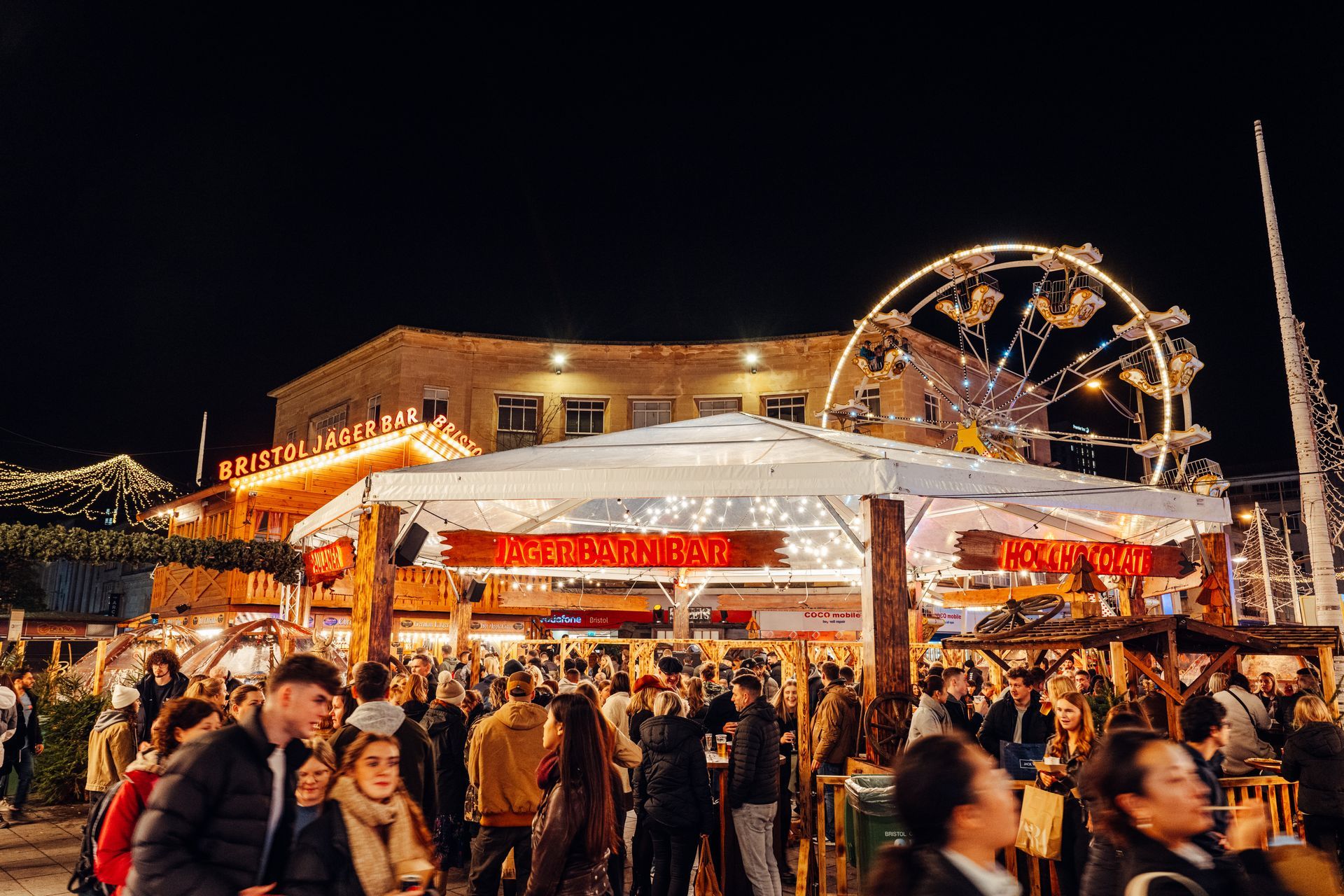 A crowd of people at a christmas market with a ferris wheel in the background