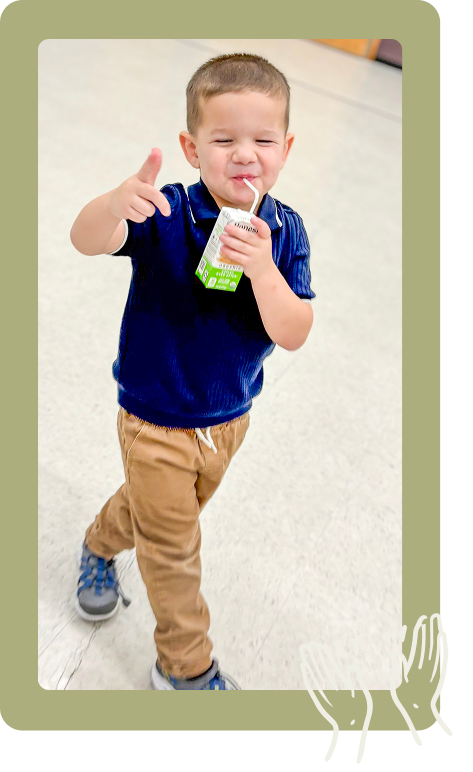 Child in blue shirt and tan pants holding a drink and giving a thumbs-up indoors.