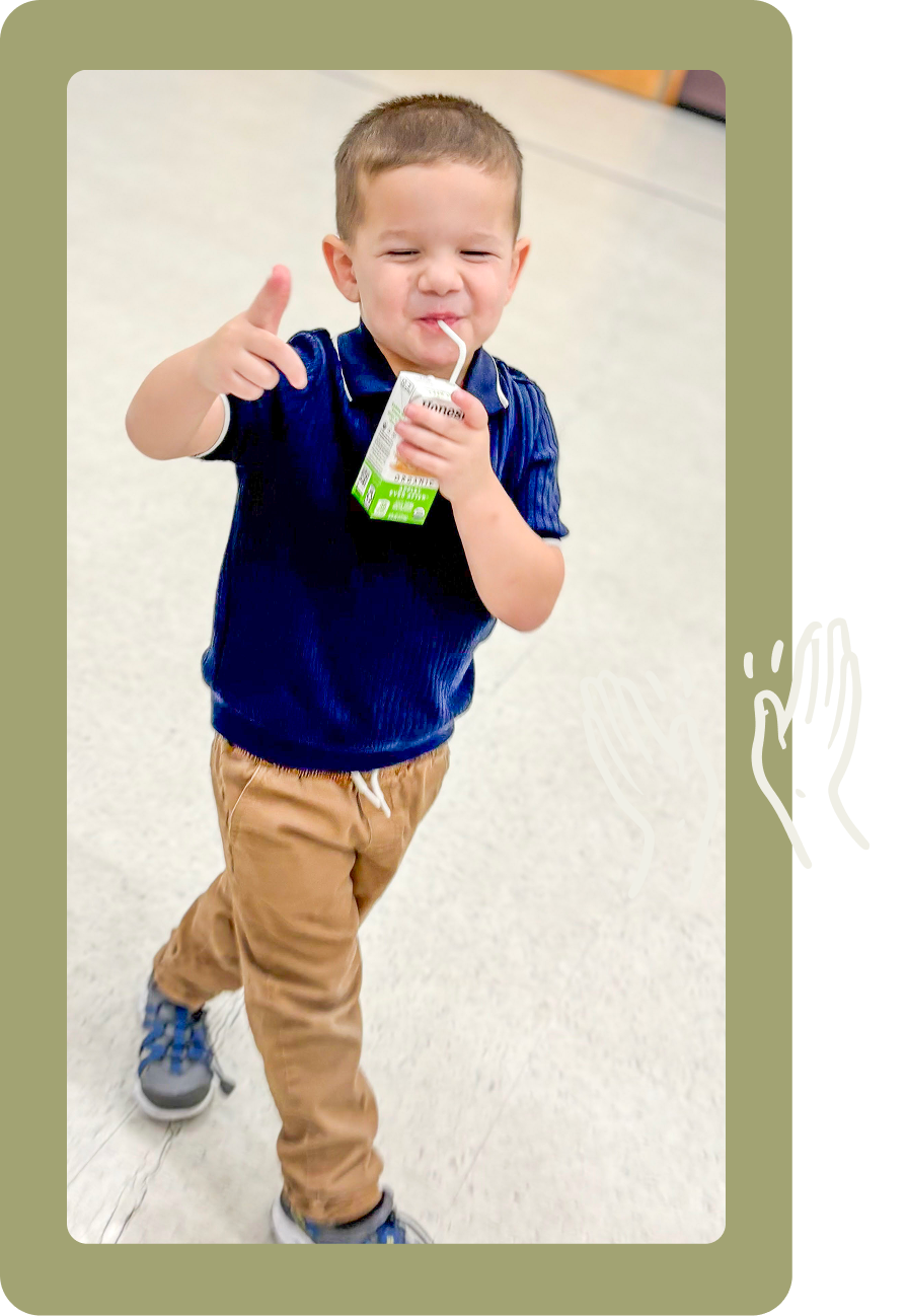 Young child in a blue shirt and tan pants walking and drinking from a juice pouch indoors.